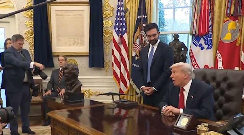 Donald Trump seated at the Resolute Desk in the Oval Office, engaging with staff and surrounded by historical artifacts and flags.