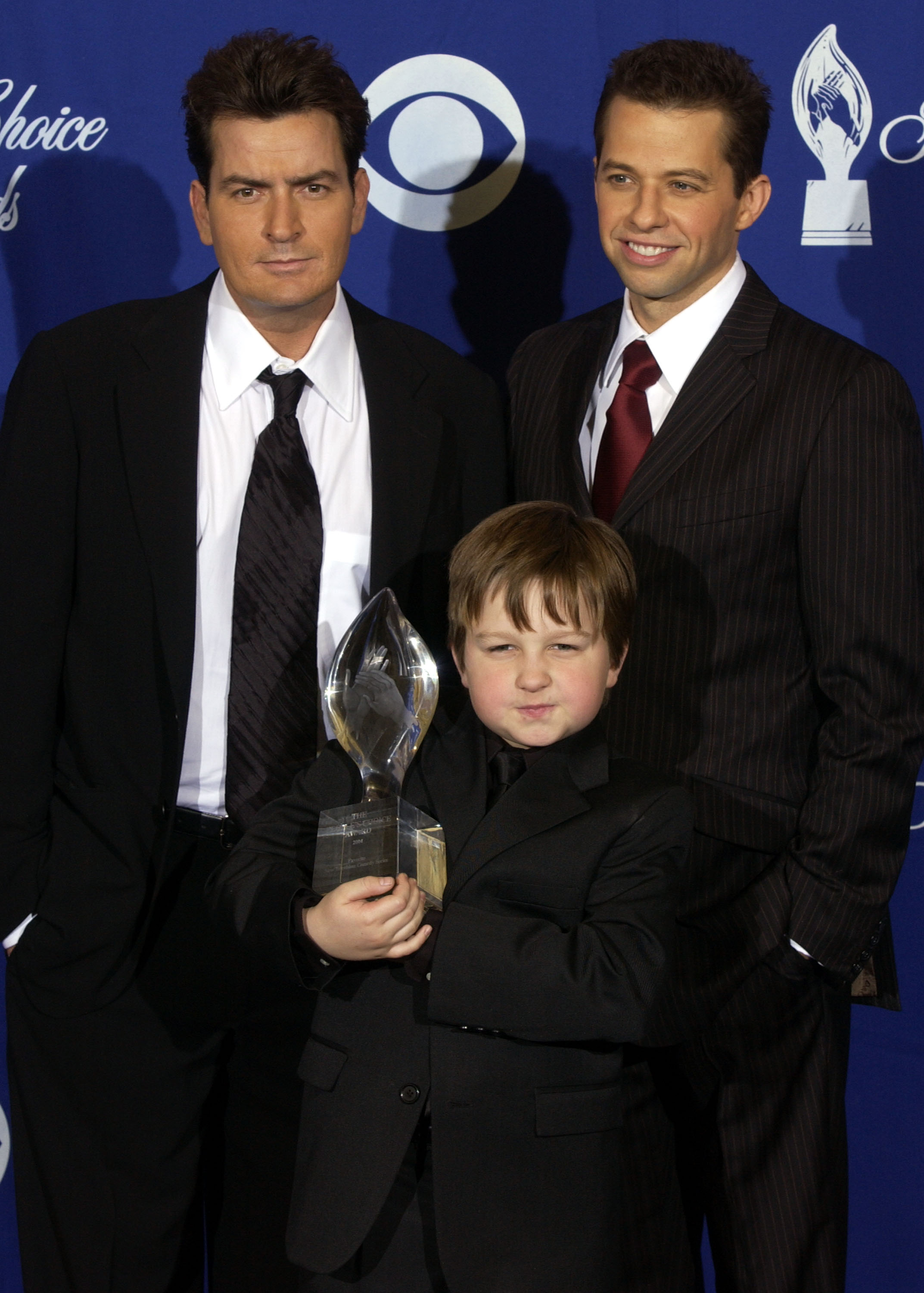 Two adult men in dark suits standing behind a young boy in a black suit holding a crystal trophy at the People's Choice Awards pressroom.