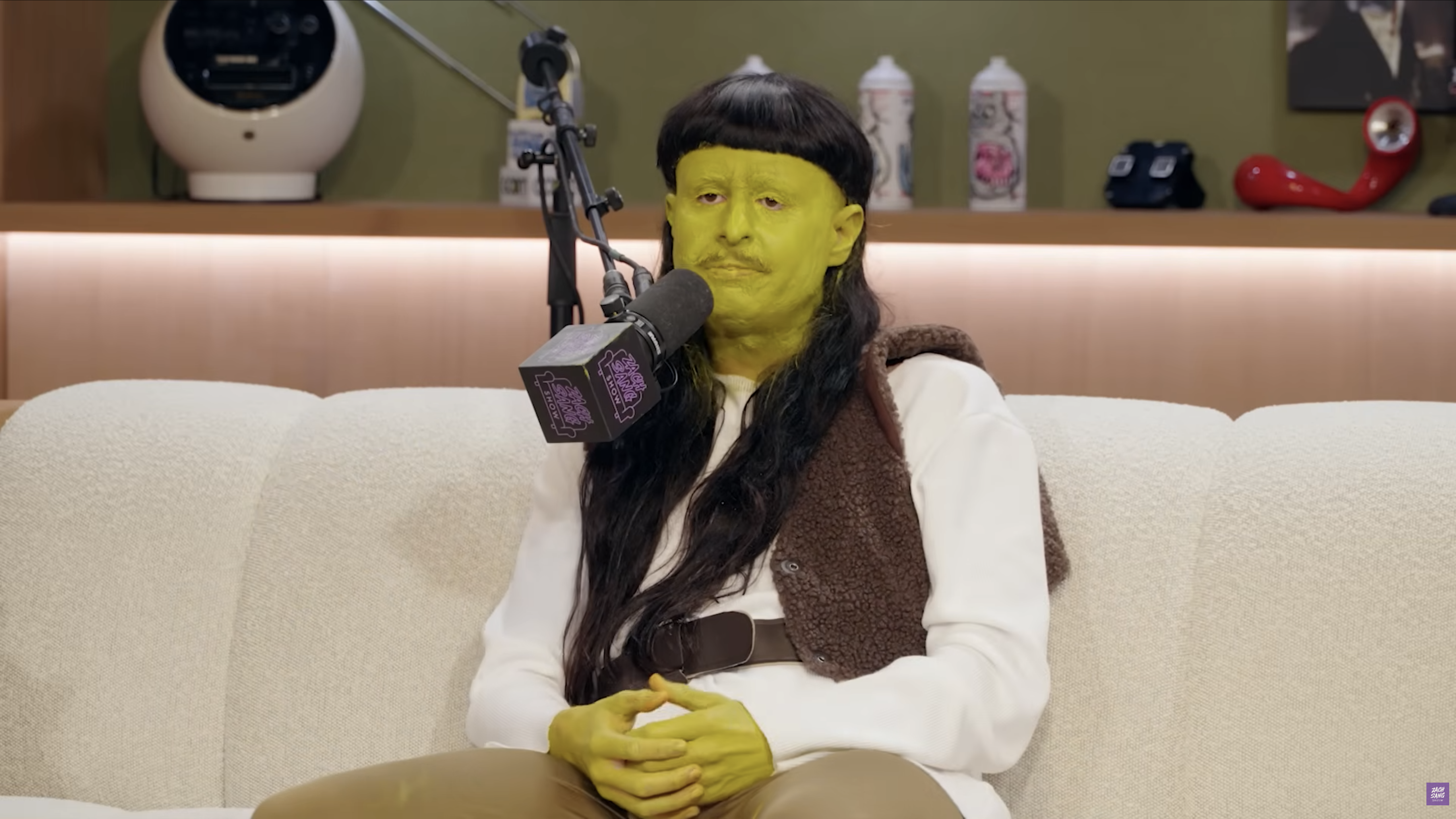 Person with green face paint, long dark wig, and brown vest seated on a white couch next to a branded podcast microphone.