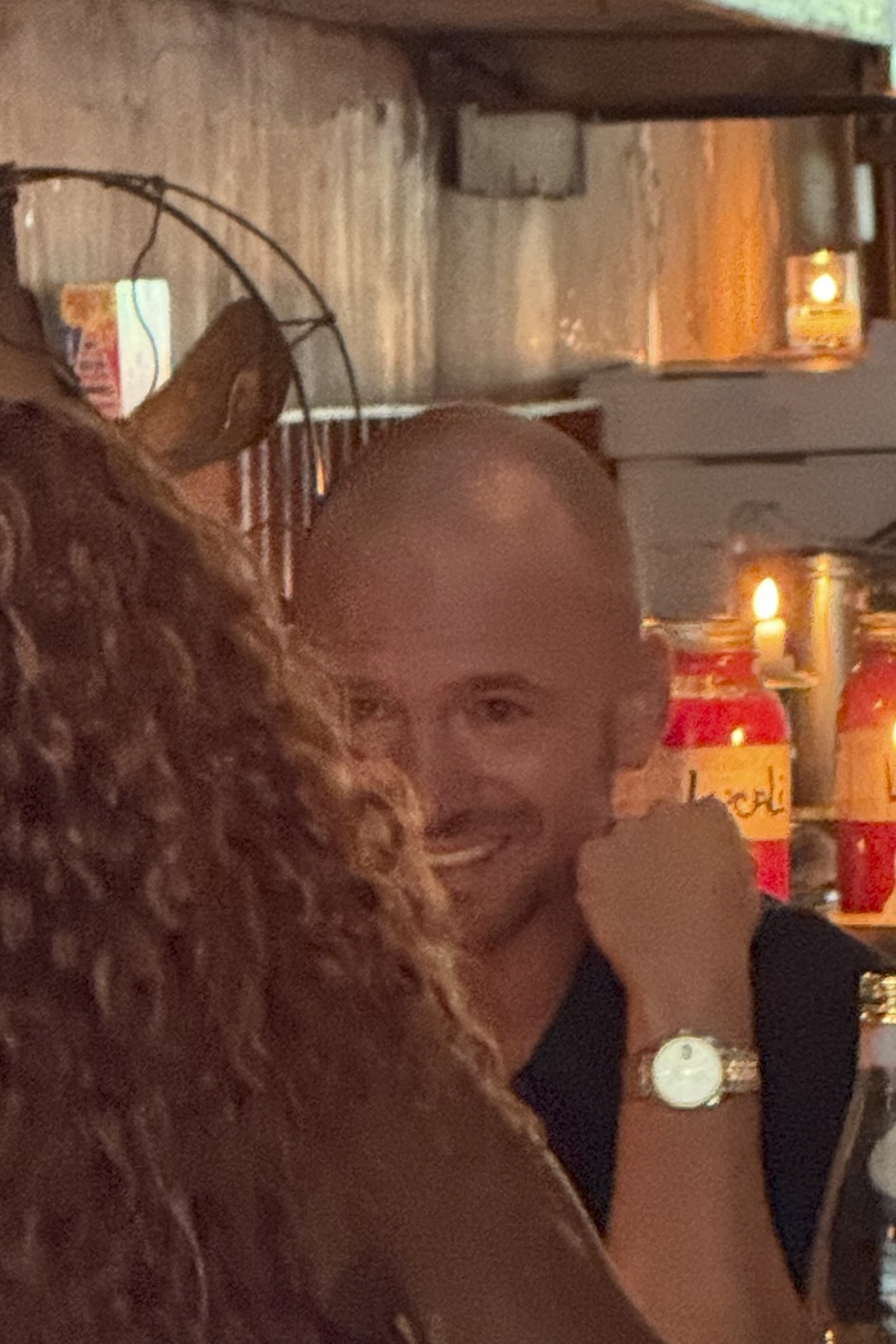 A smiling bald man seen over the shoulder of a curly-haired person at a warmly lit restaurant table with candles and red drinks in the background.