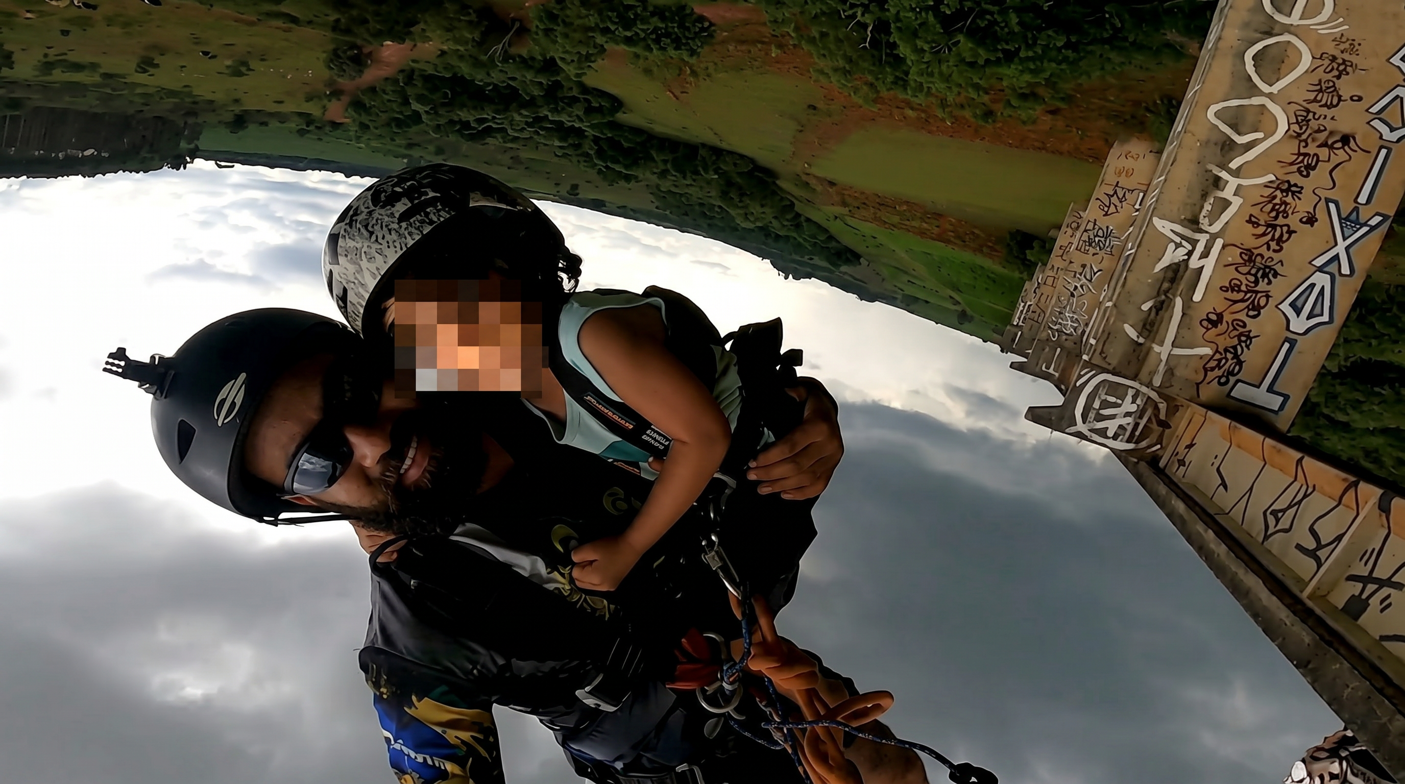 Upside-down GoPro selfie of a helmeted man in sunglasses holding a harnessed child during a jump from a graffiti-covered bridge structure.