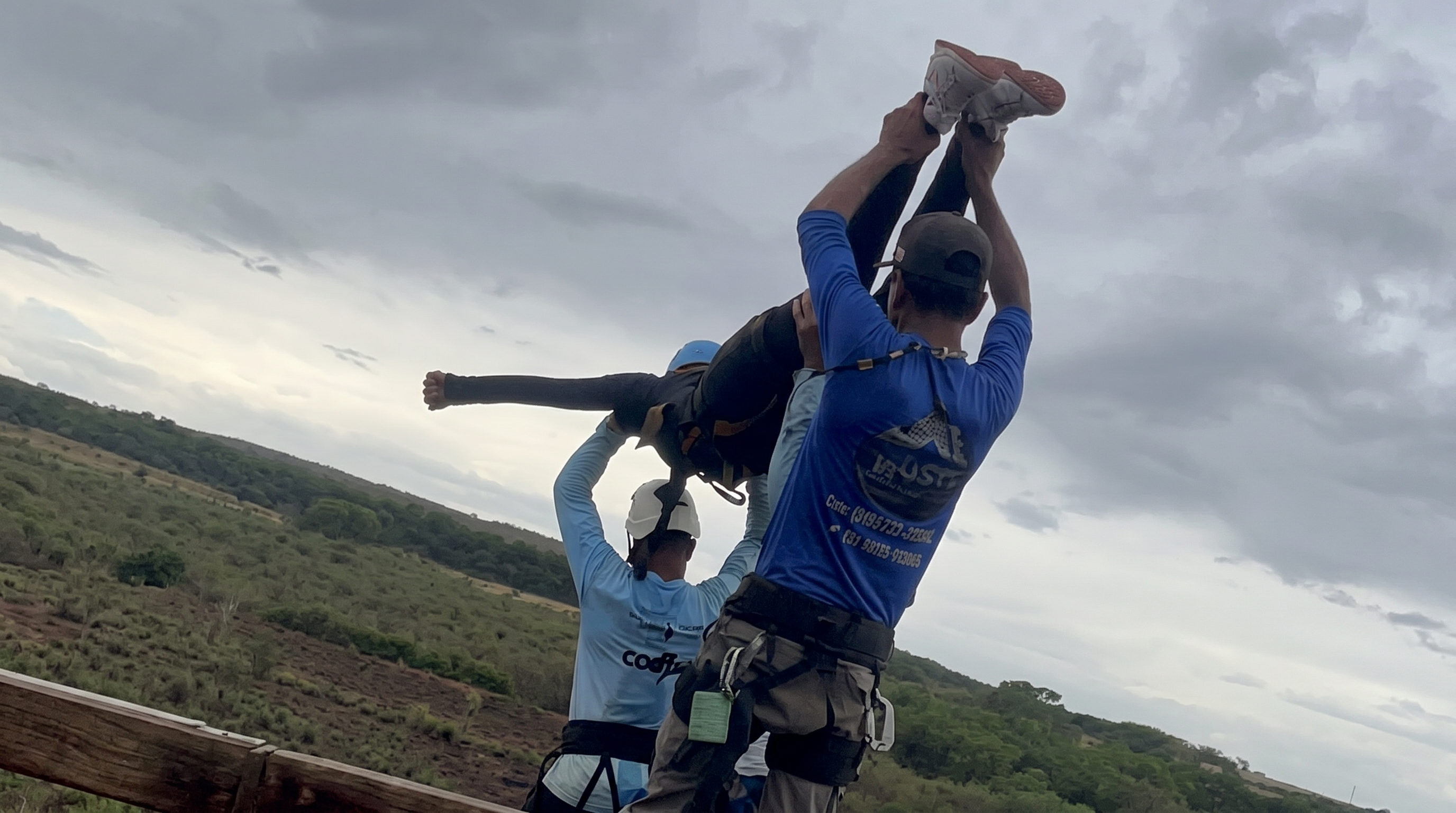 Two men in blue shirts and harnesses hold a harnessed person upside-down by the legs at the edge of a jump platform, with green hills in the background.