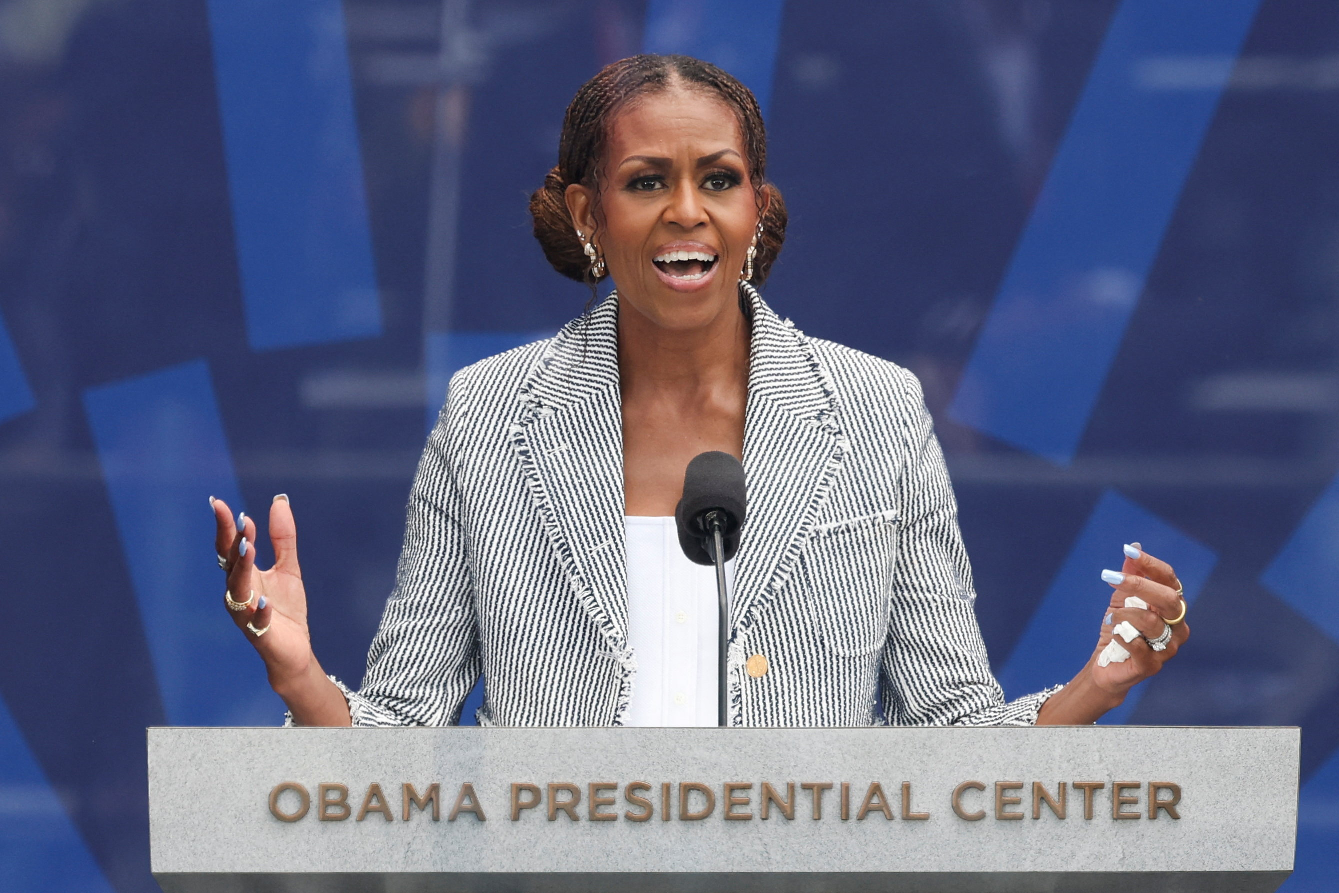 Michelle Obama speaks passionately at a podium labeled 'Obama Presidential Center,' gesturing with both hands, wearing a striped blazer.