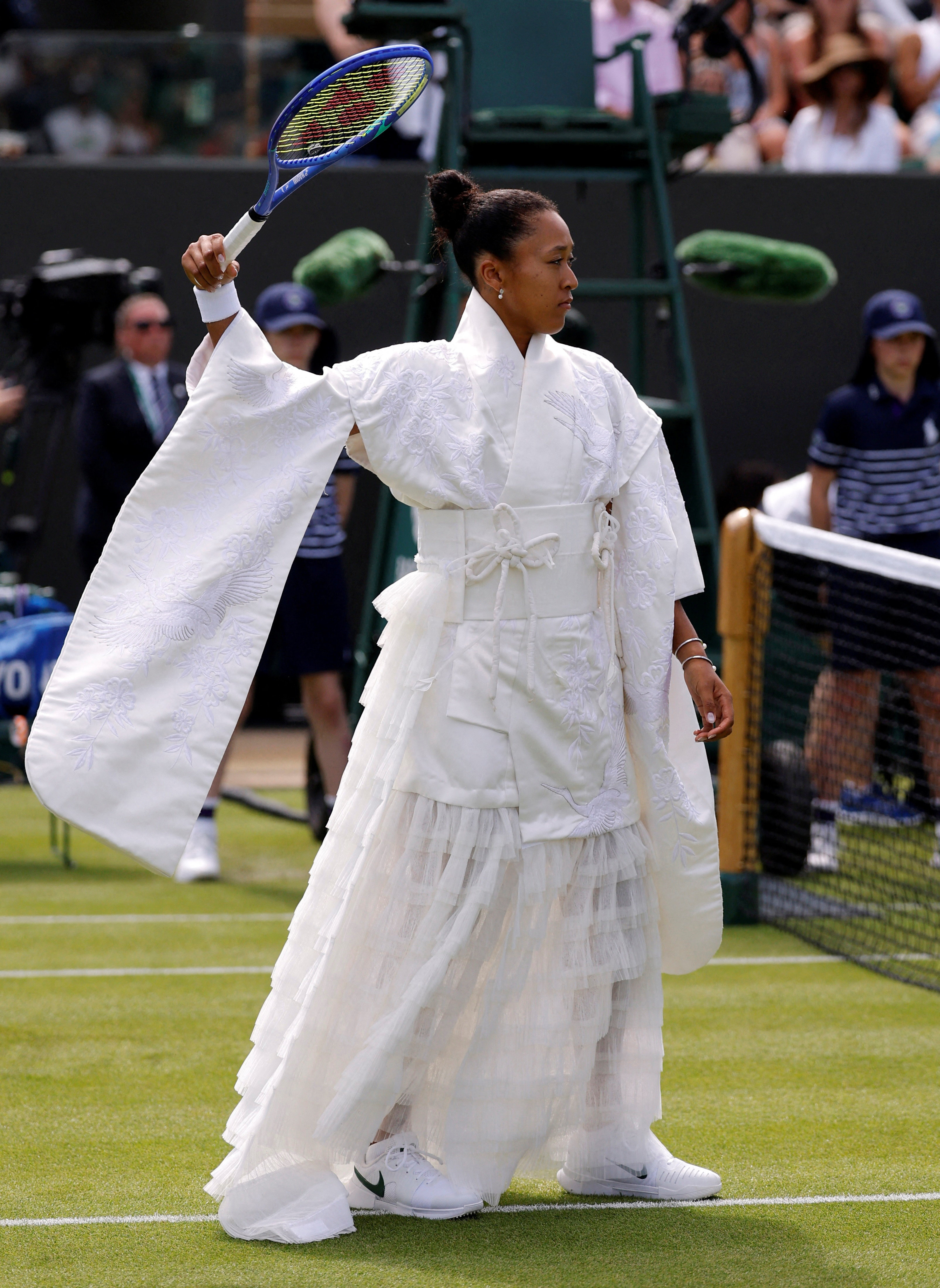 Tennis player in a flowing white embroidered kimono-style gown raises a blue Yonex racket on a Wimbledon grass court.