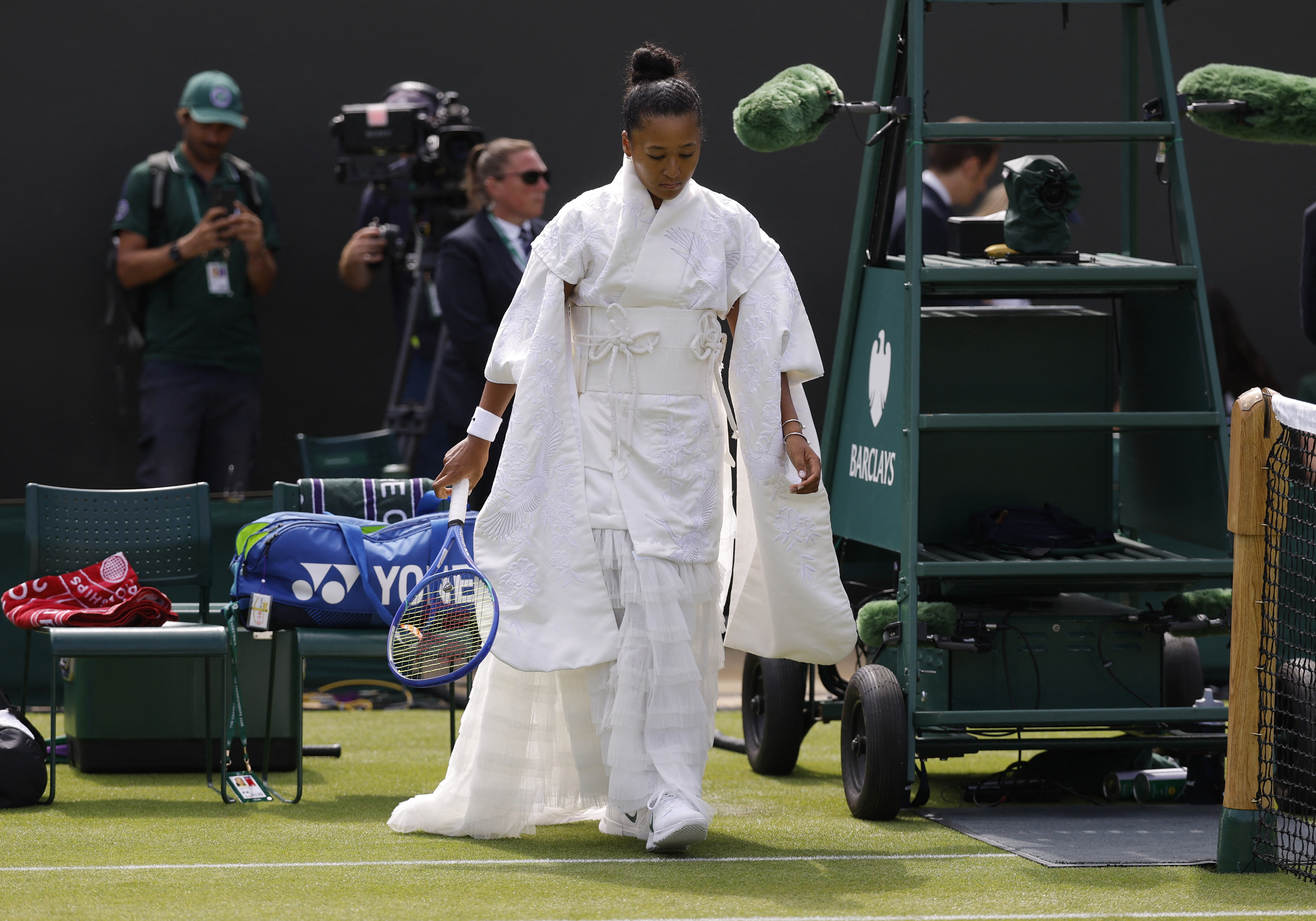 Tennis player in a white kimono-inspired gown carrying a blue Yonex racket walks near the umpire's chair on a Wimbledon grass court, with cameramen in the background.