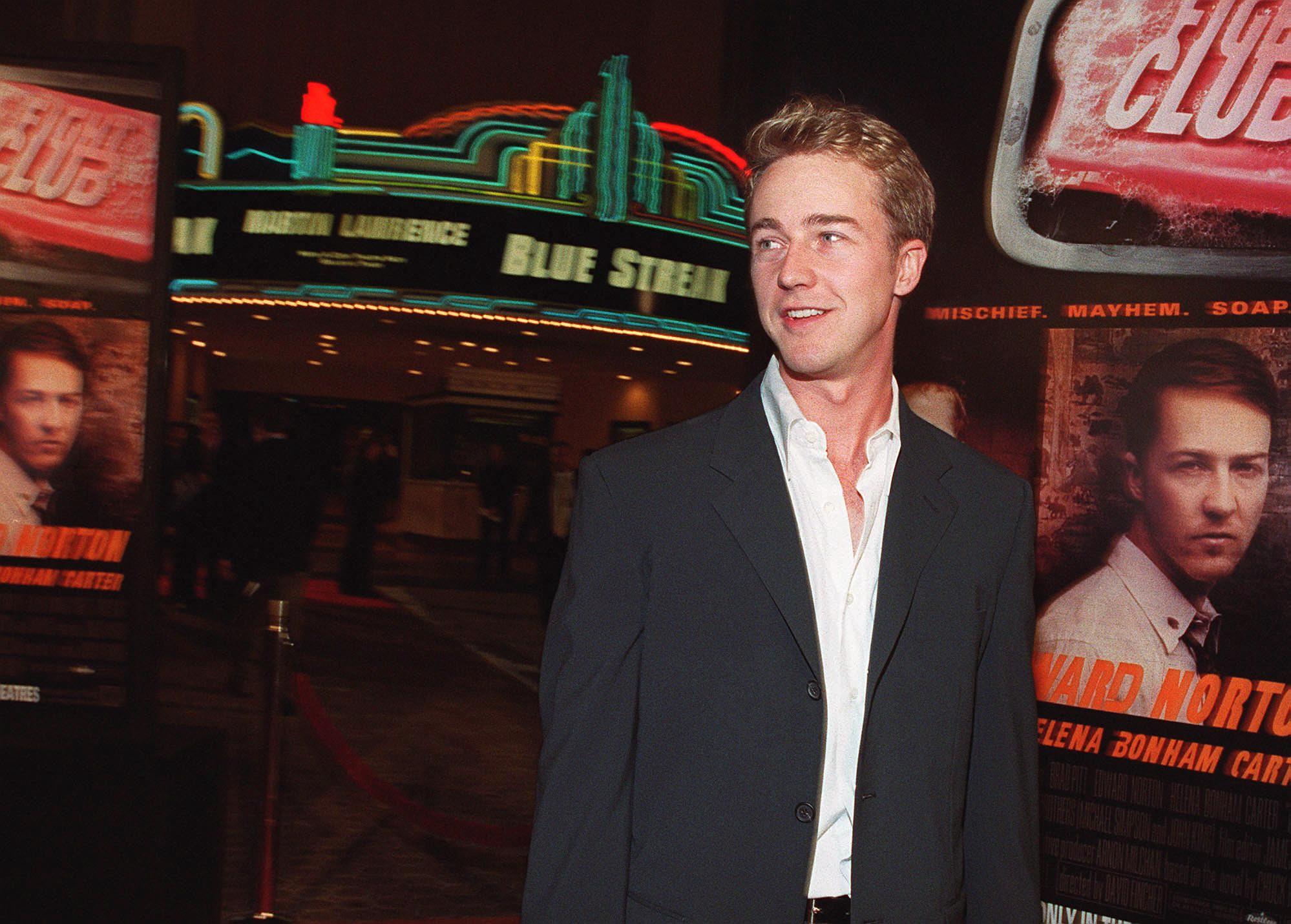 Edward Norton in a dark blazer and open white shirt standing in front of Fight Club movie posters at a theater premiere.