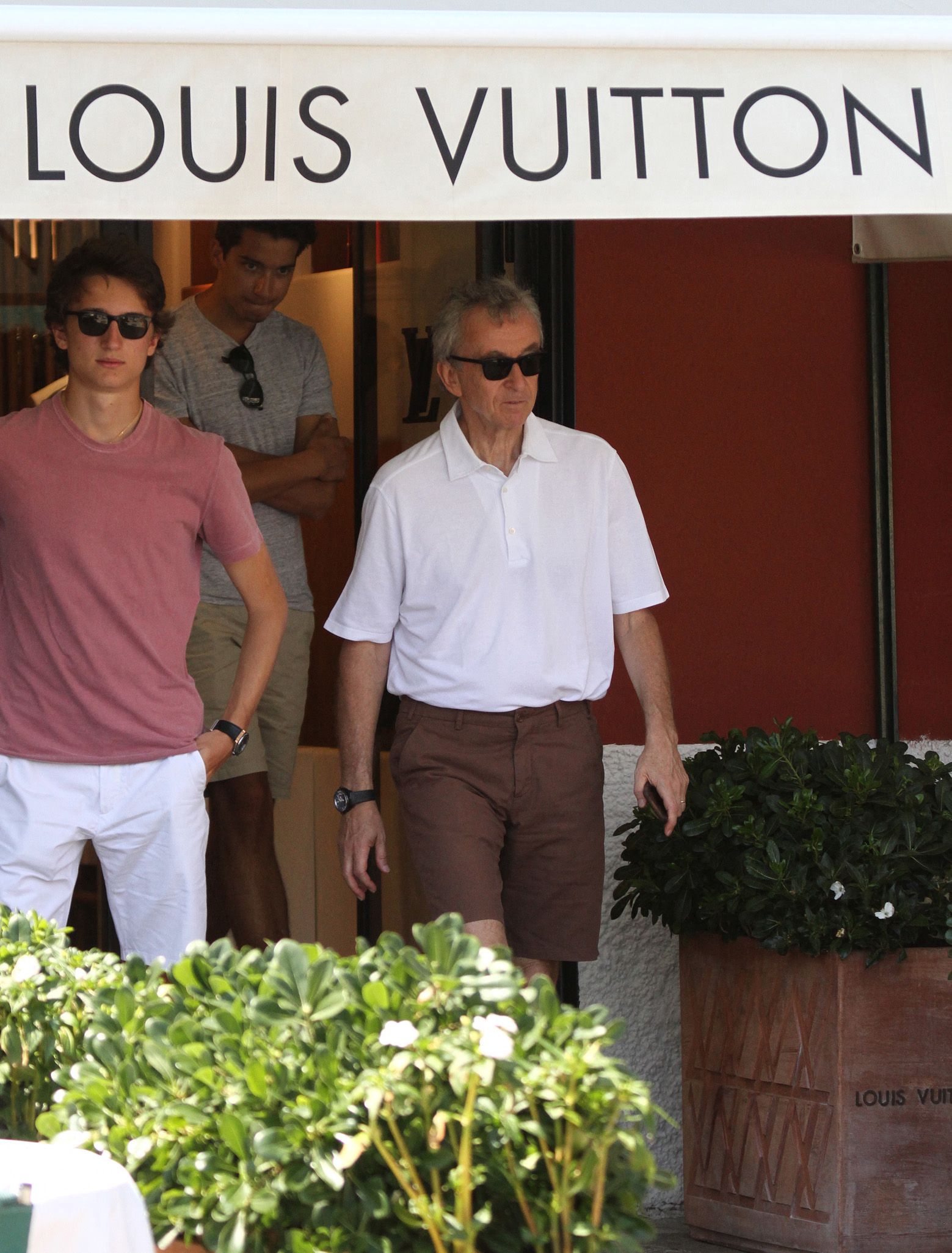 An older man in a white polo and brown shorts and a younger man in a pink t-shirt walk out of a Louis Vuitton store entrance.