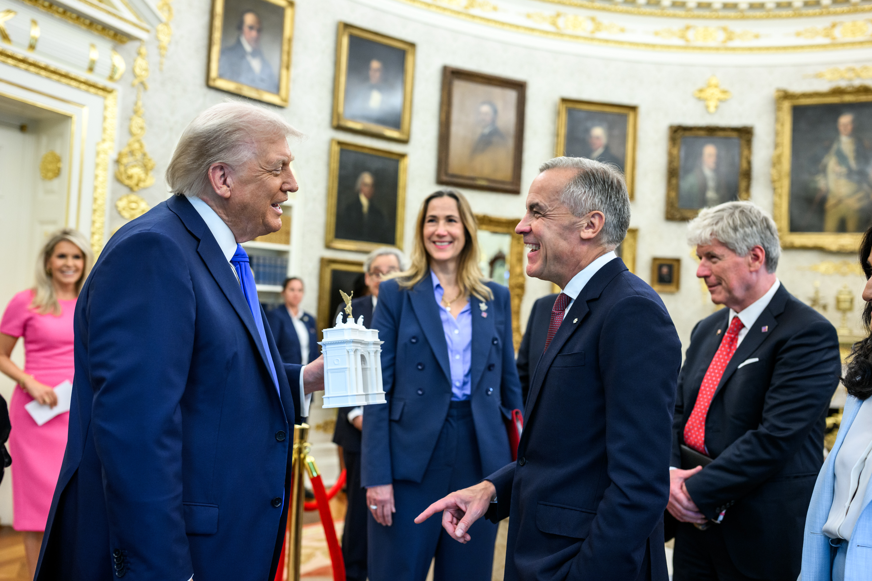 President Trump holding a white miniature model of a triumphal arch while speaking with Canadian PM Mark Carney inside the White House, surrounded by aides and portrait paintings.