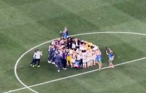 A diverse group of soccer players and staff huddle together on the field, celebrating a significant victory after a match.