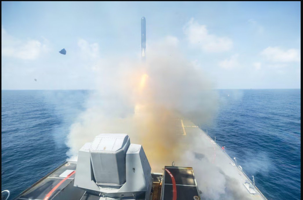Missile launch from a naval vessel over the ocean, creating smoke and water spray against a clear blue sky.