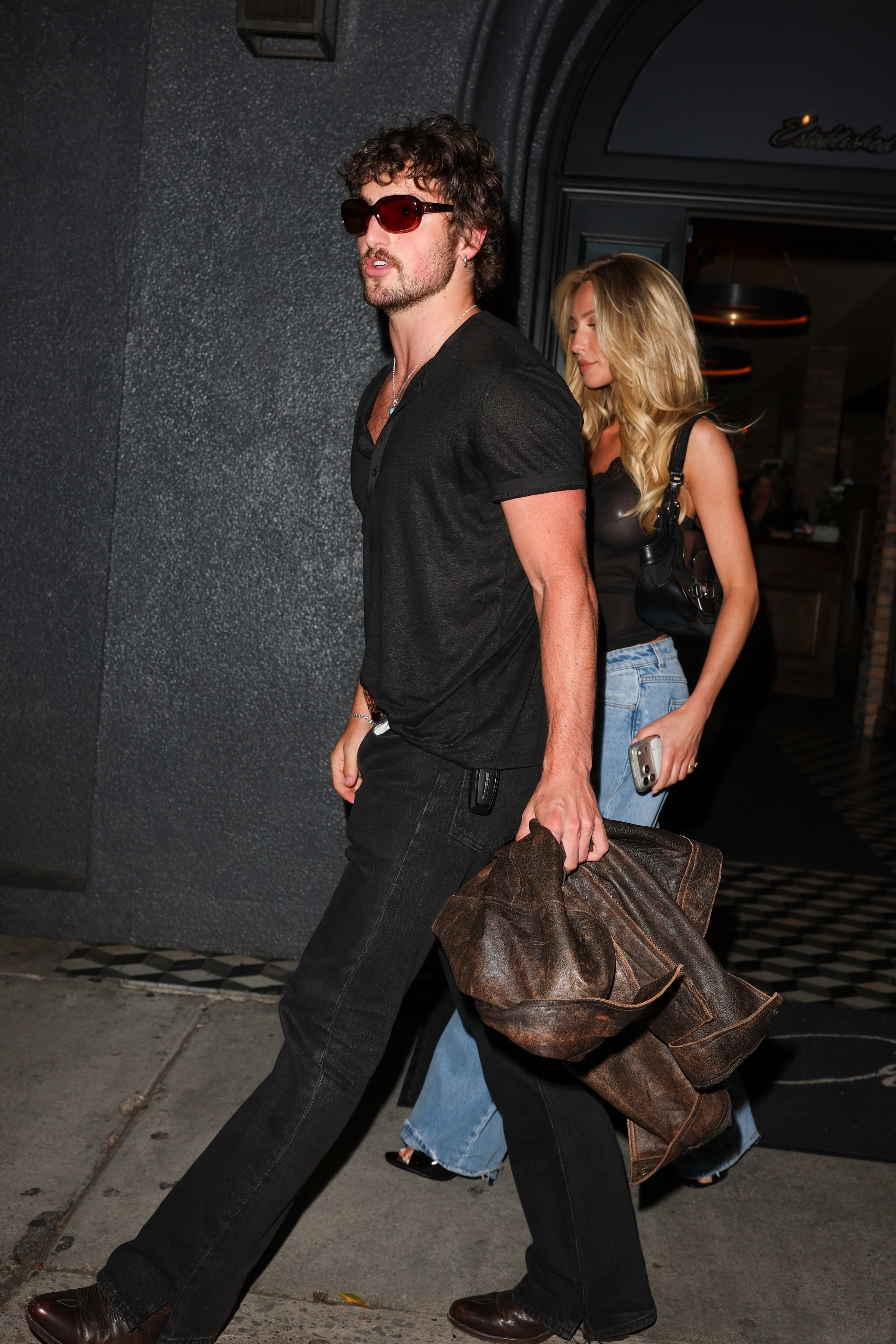 A curly-haired man in a black t-shirt and dark jeans carries a large brown leather bag while a blonde woman in blue jeans walks behind him outside a building at night.