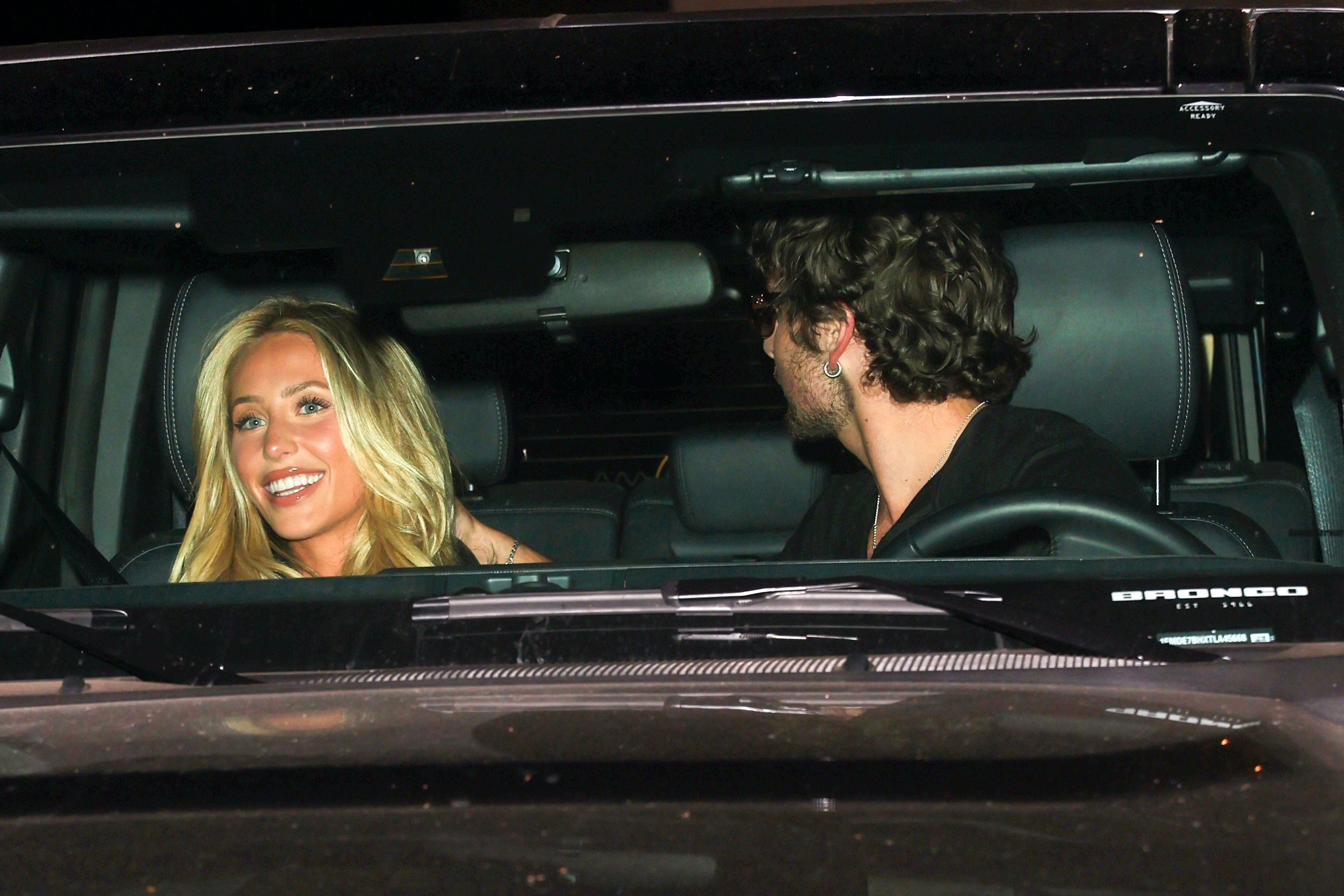 Smiling blonde woman and curly-haired man sitting inside a black Ford Bronco at night.