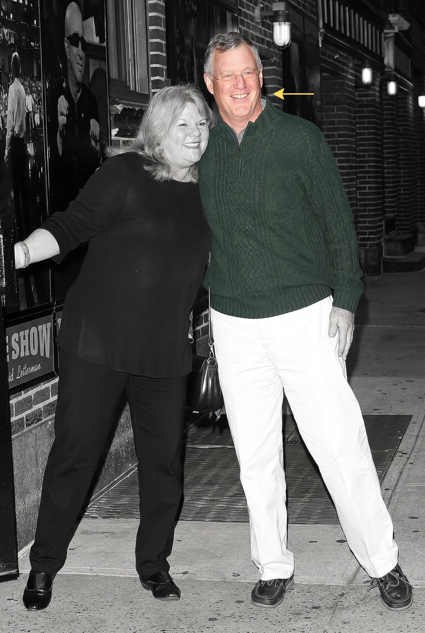 A man in a green sweater and white pants and a woman in all black smile together outside the Late Show with David Letterman, in a black-and-white photo with the man's sweater colorized green.
