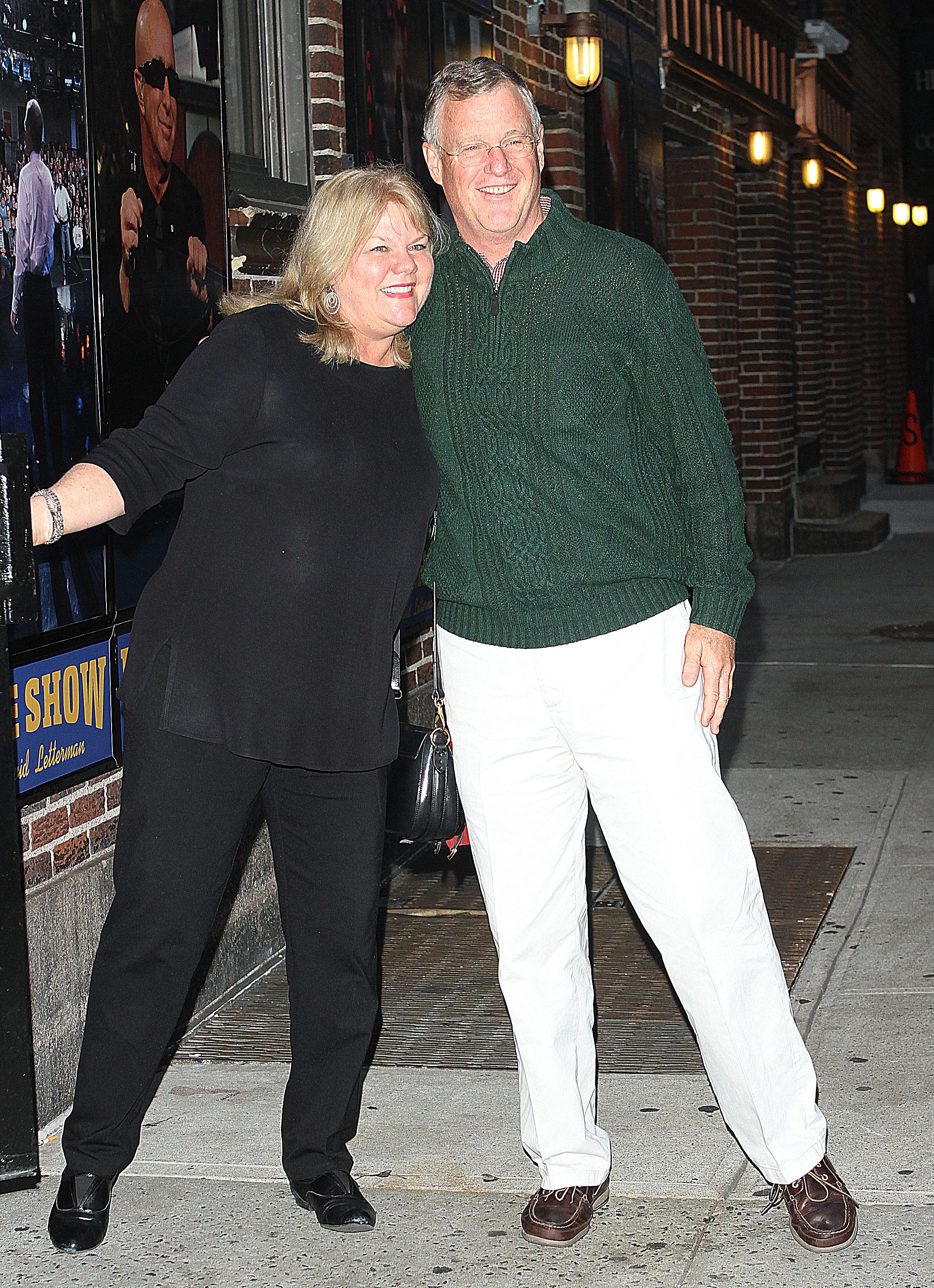A smiling couple — a woman in black and a tall man in a green cable-knit sweater and white pants — pose together on a sidewalk outside the Late Show with David Letterman.