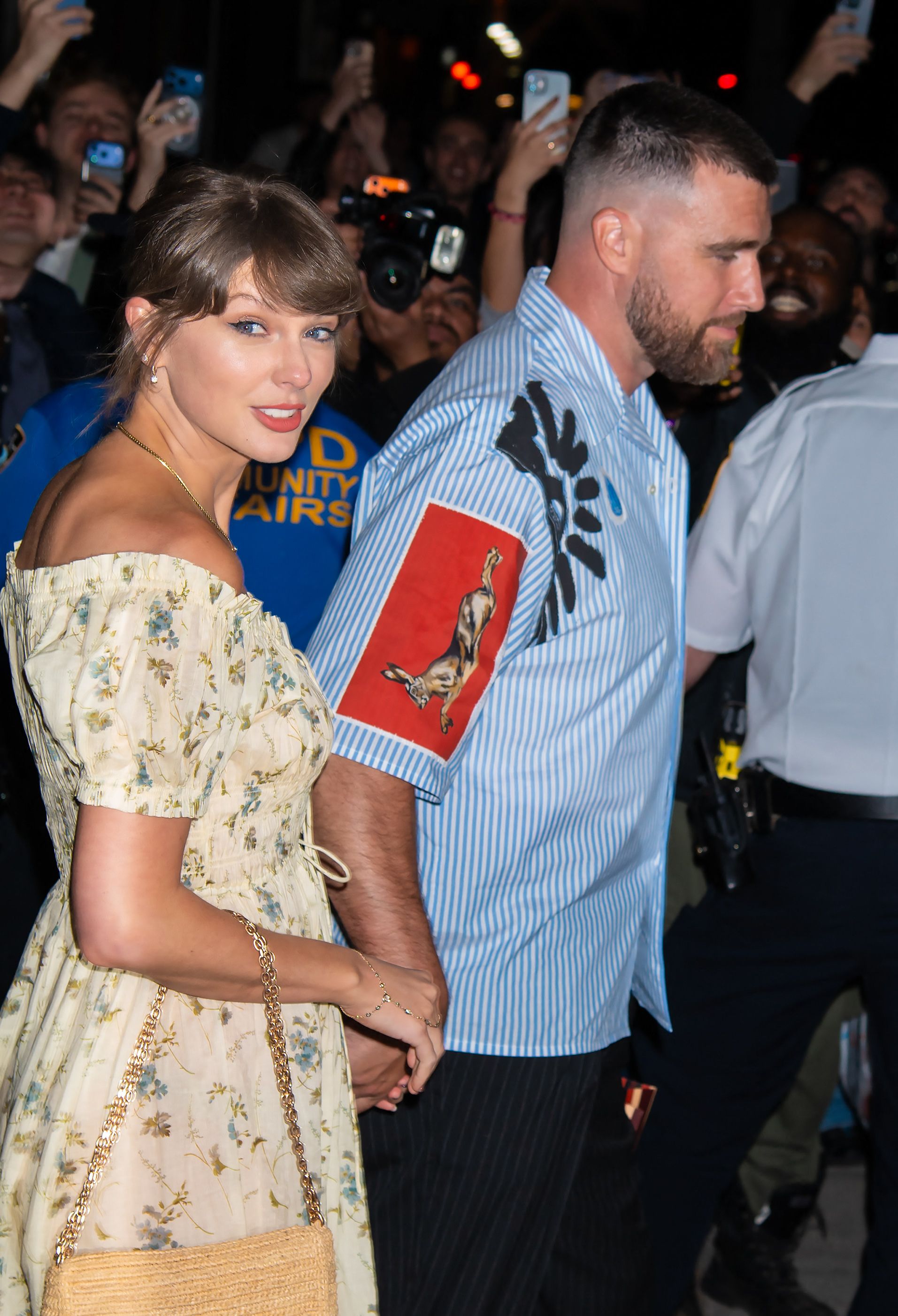 A woman in a cream floral off-shoulder dress and a tall bearded man in a blue striped shirt walk hand-in-hand through a crowd of fans and photographers at night.