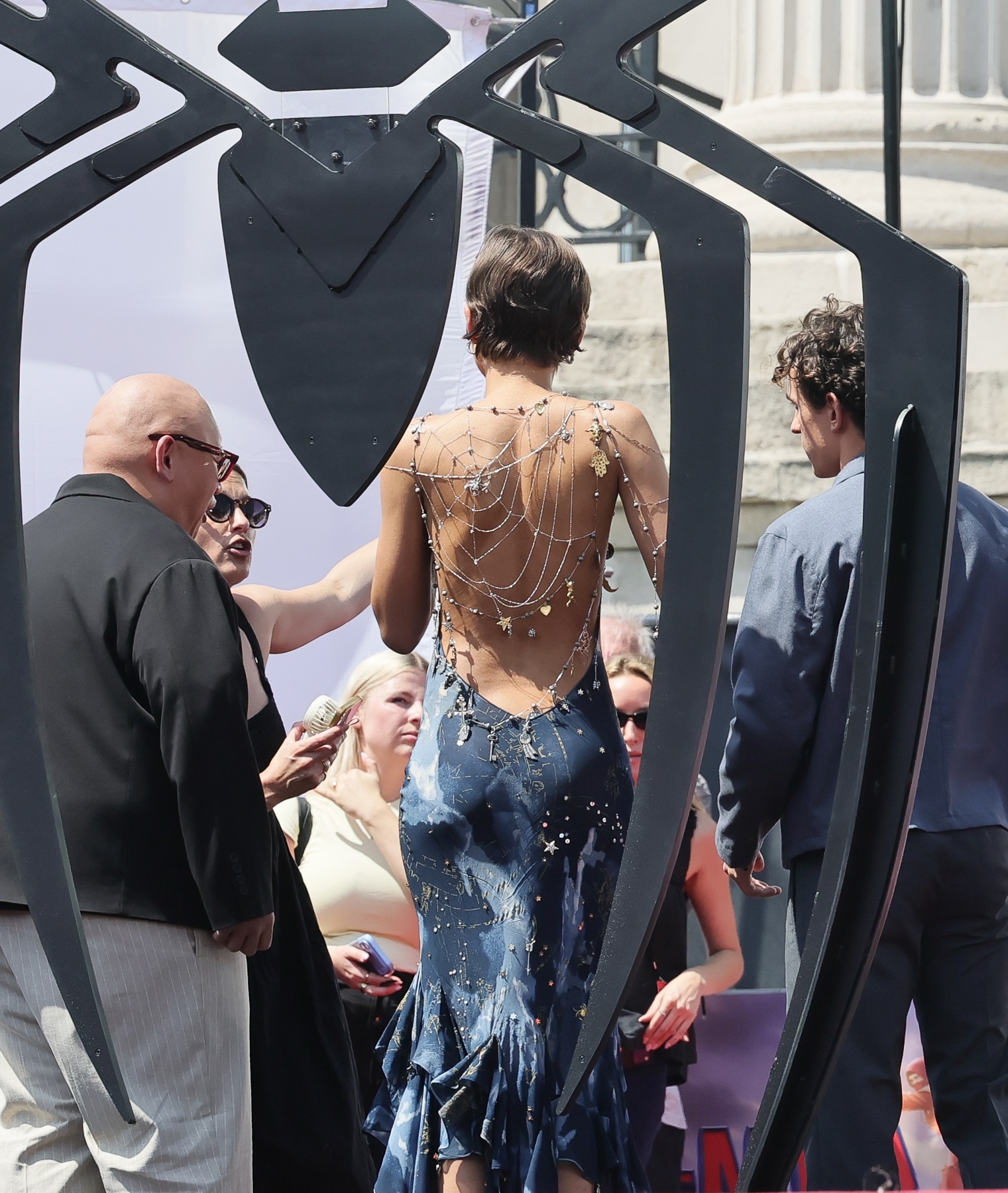 Back view of woman in backless blue dress with intricate silver spider web chain body jewelry covering her entire back, standing near a large black spider-shaped structure.