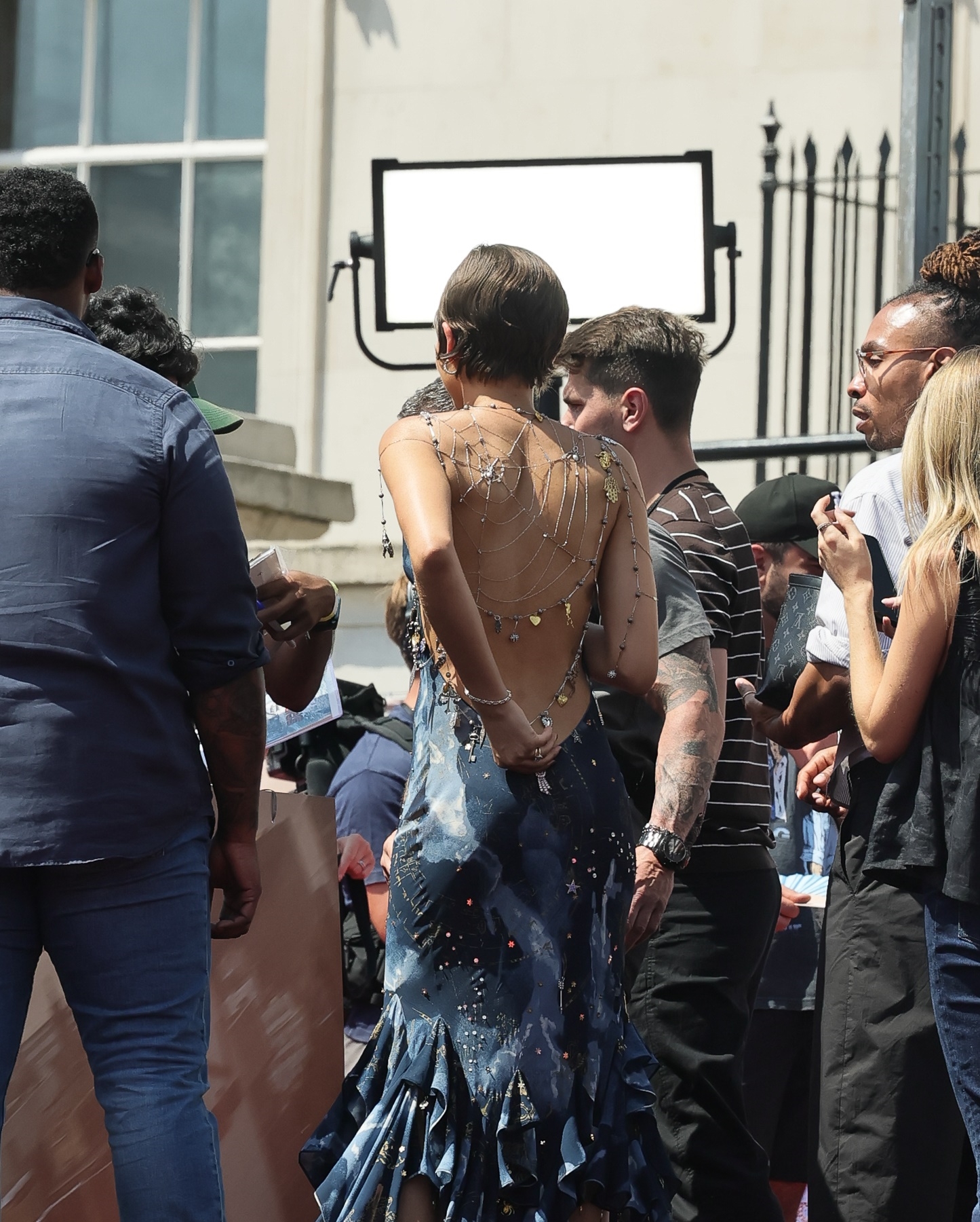 Back view of woman in backless blue ruffle-hem dress with silver spider web chain jewelry draped across her back, standing amid a crowd on a red carpet.