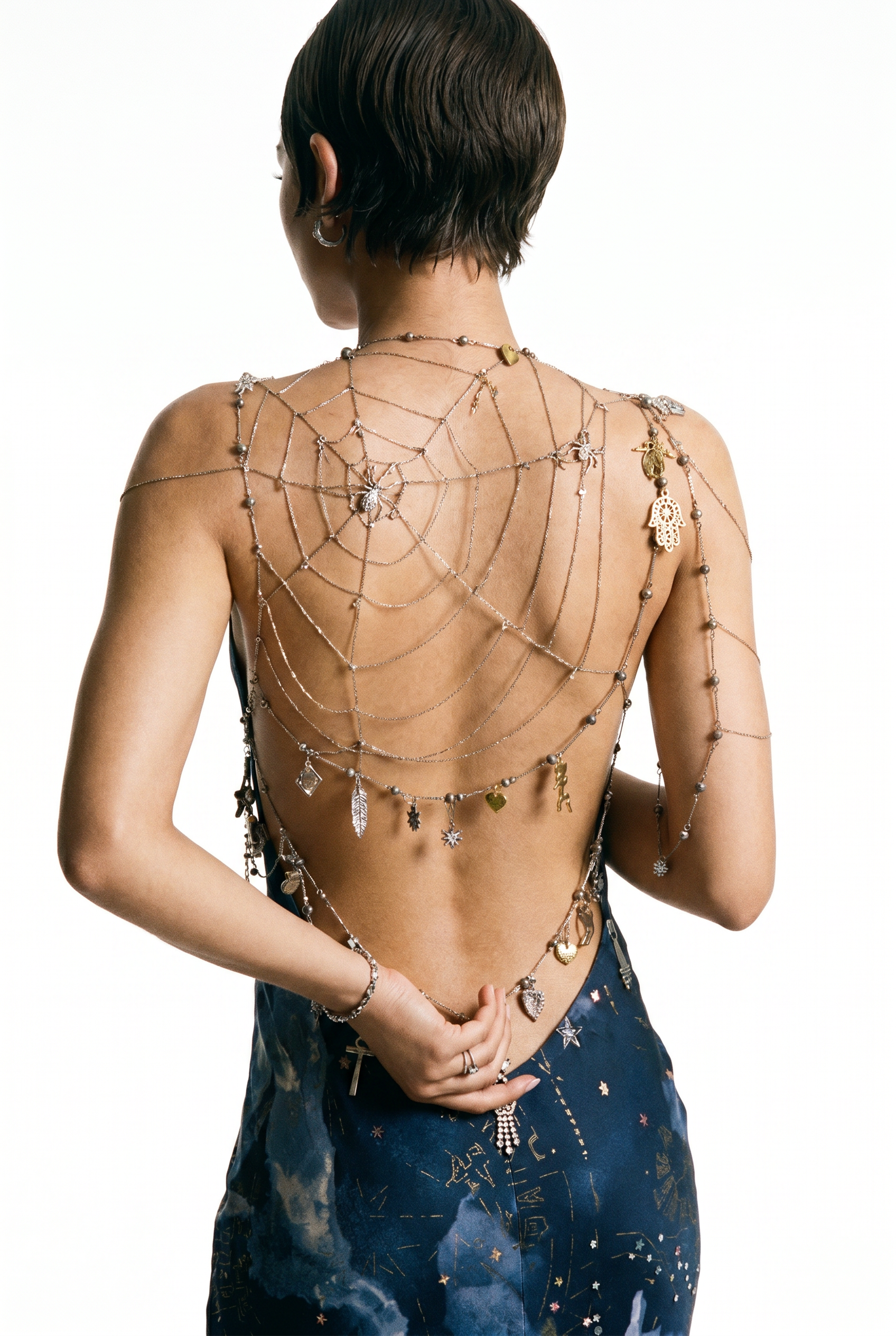 Close-up back view of woman in backless blue celestial-print dress wearing an elaborate silver chain spider web body jewelry piece with gold and silver charms.