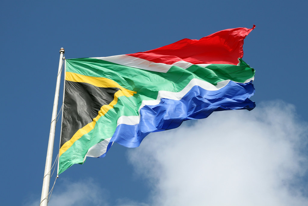South African flag waving against a clear blue sky, showcasing vibrant colors of green, black, yellow, red, and blue.