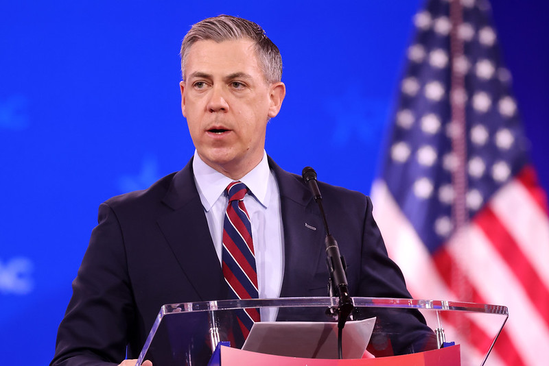 Political speaker at a podium addressing an audience, with an American flag in the background, during a conference event.