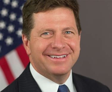 Professional headshot of a smiling man in a suit with a blue tie, featuring the American flag in the background.