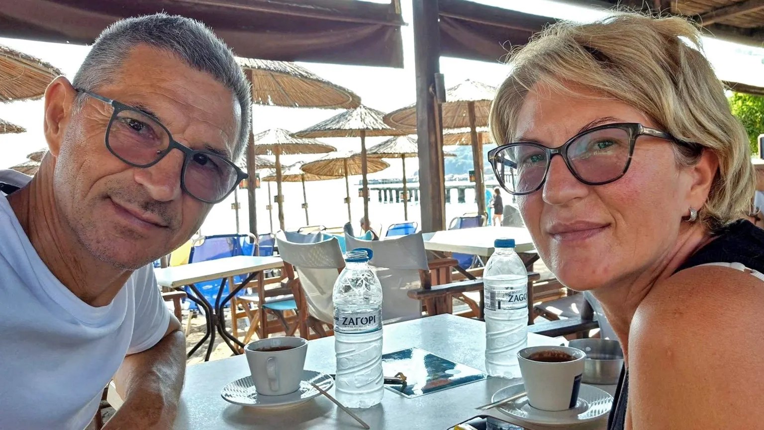 A man and woman wearing glasses smile at the camera while seated at an outdoor beach café table with two water bottles and coffee cups.