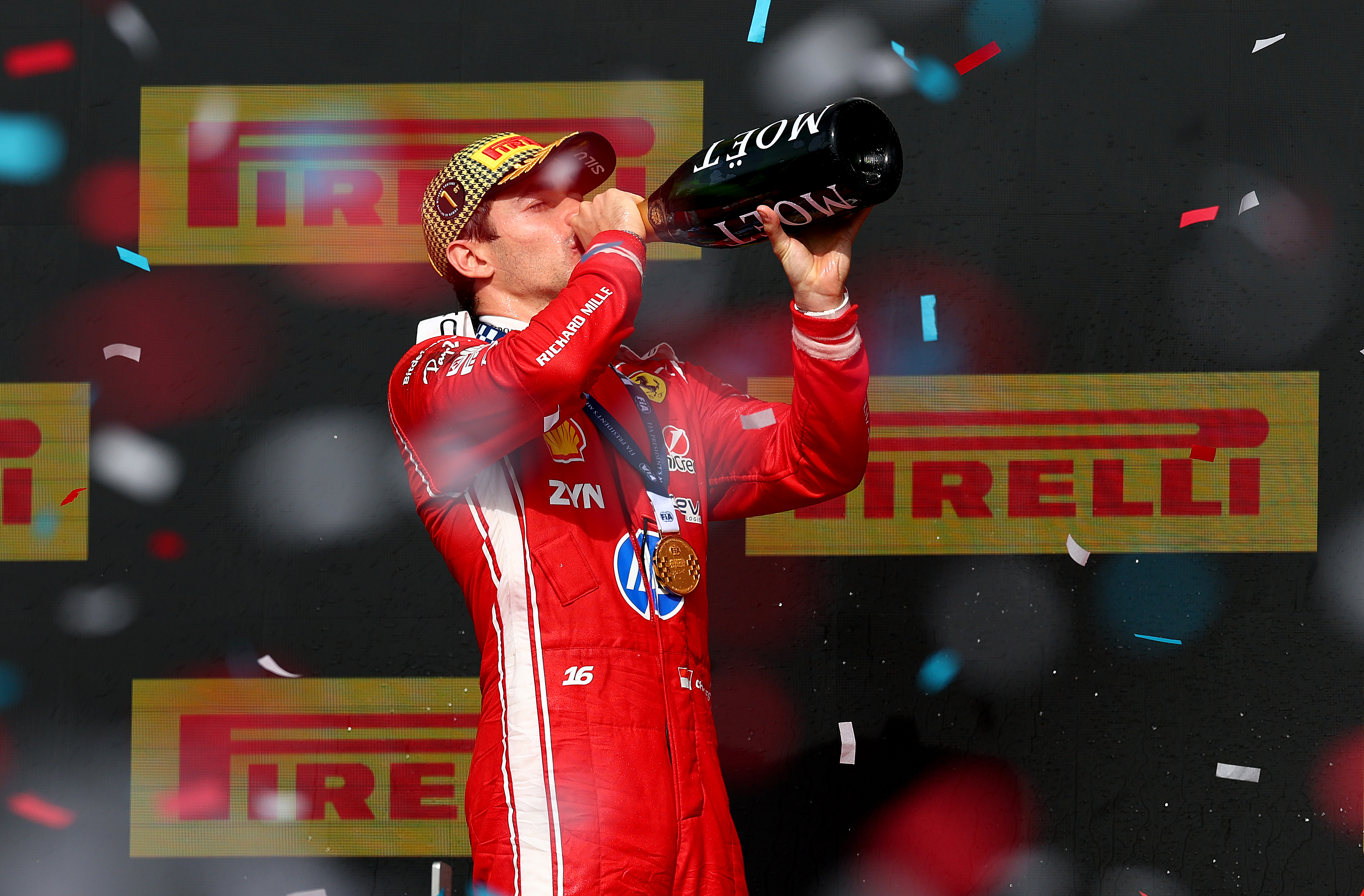 Ferrari driver Charles Leclerc in red race suit drinking from a Moët champagne bottle on the podium surrounded by confetti.