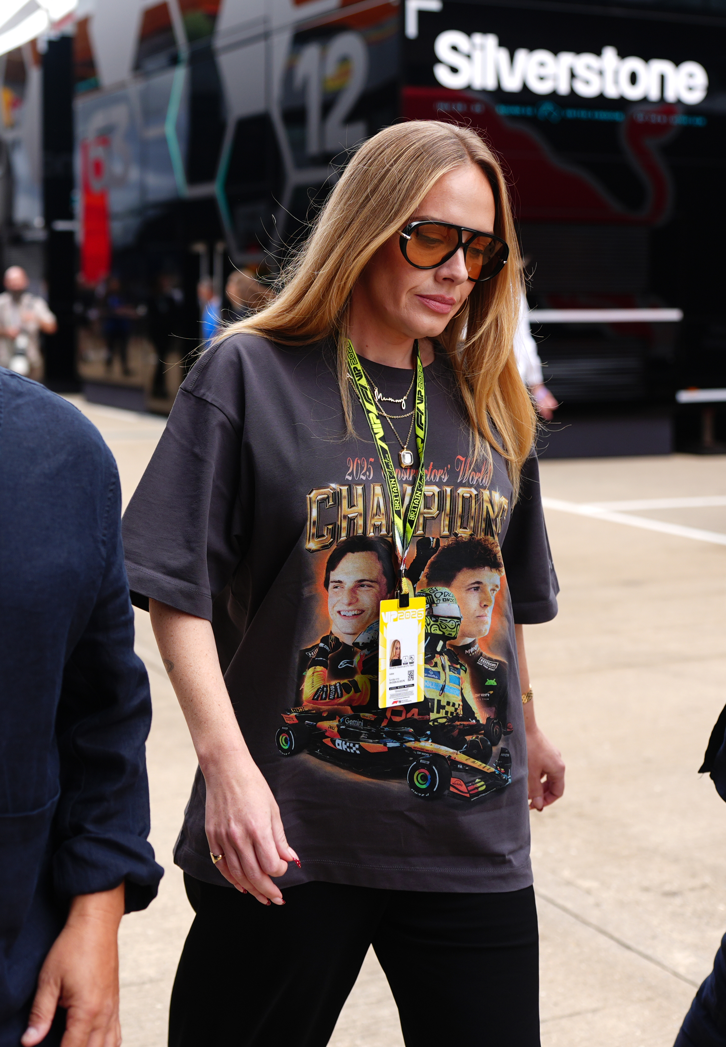 Adele walking in the Silverstone paddock wearing a dark McLaren champions graphic tee, amber-tinted sunglasses, and a green VIP lanyard.
