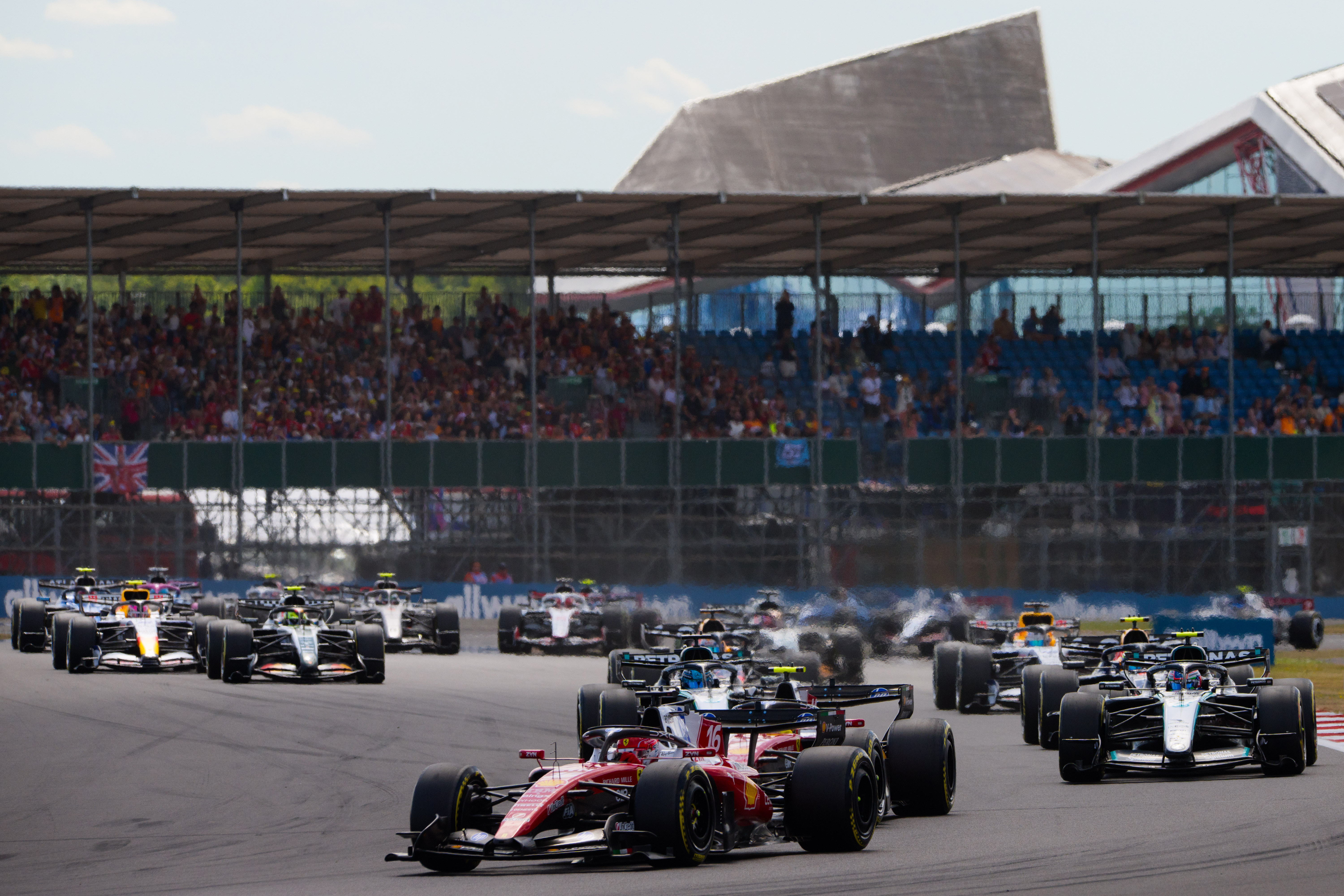 Ferrari No. 16 car leading a field of Formula 1 cars at the start of the British Grand Prix at Silverstone, with packed grandstands in the background.
