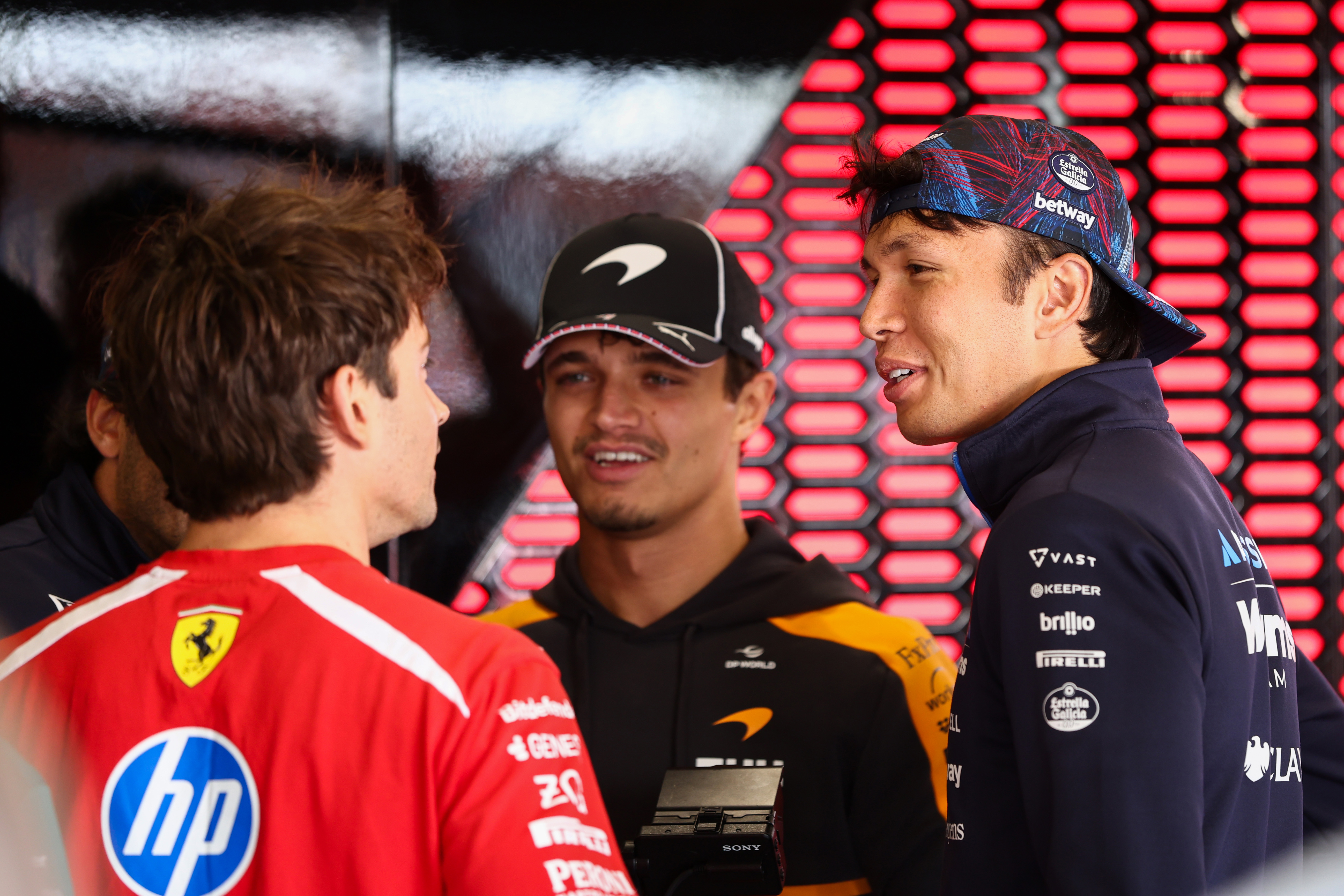Charles Leclerc in Ferrari red, Lando Norris in McLaren black, and Alexander Albon in Williams navy talk together at the Silverstone drivers parade.