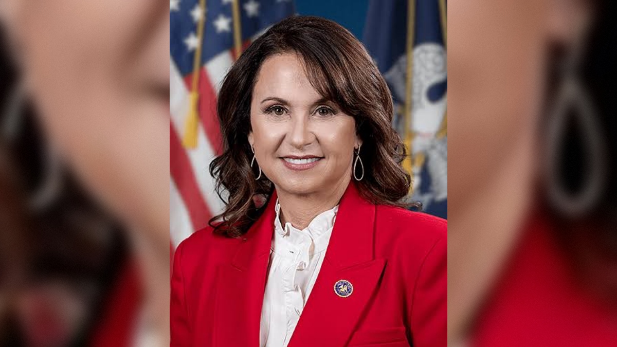 Portrait of a woman in a red blazer with a white blouse, smiling against a backdrop of flags, representing leadership and professionalism.