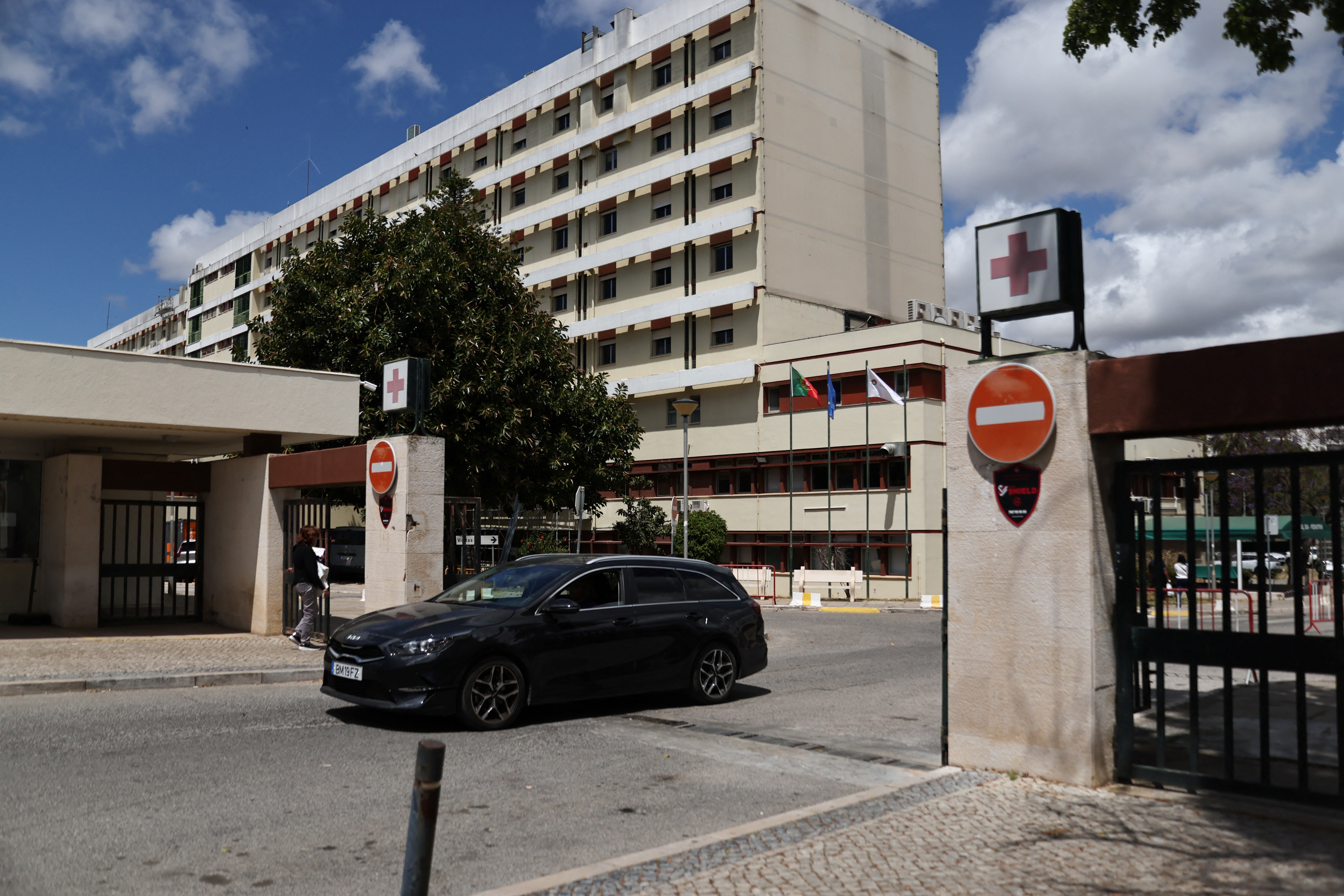Exterior of a multi-story hospital building with red cross signs at the entrance gate, a dark car passing through, under a partly cloudy sky.