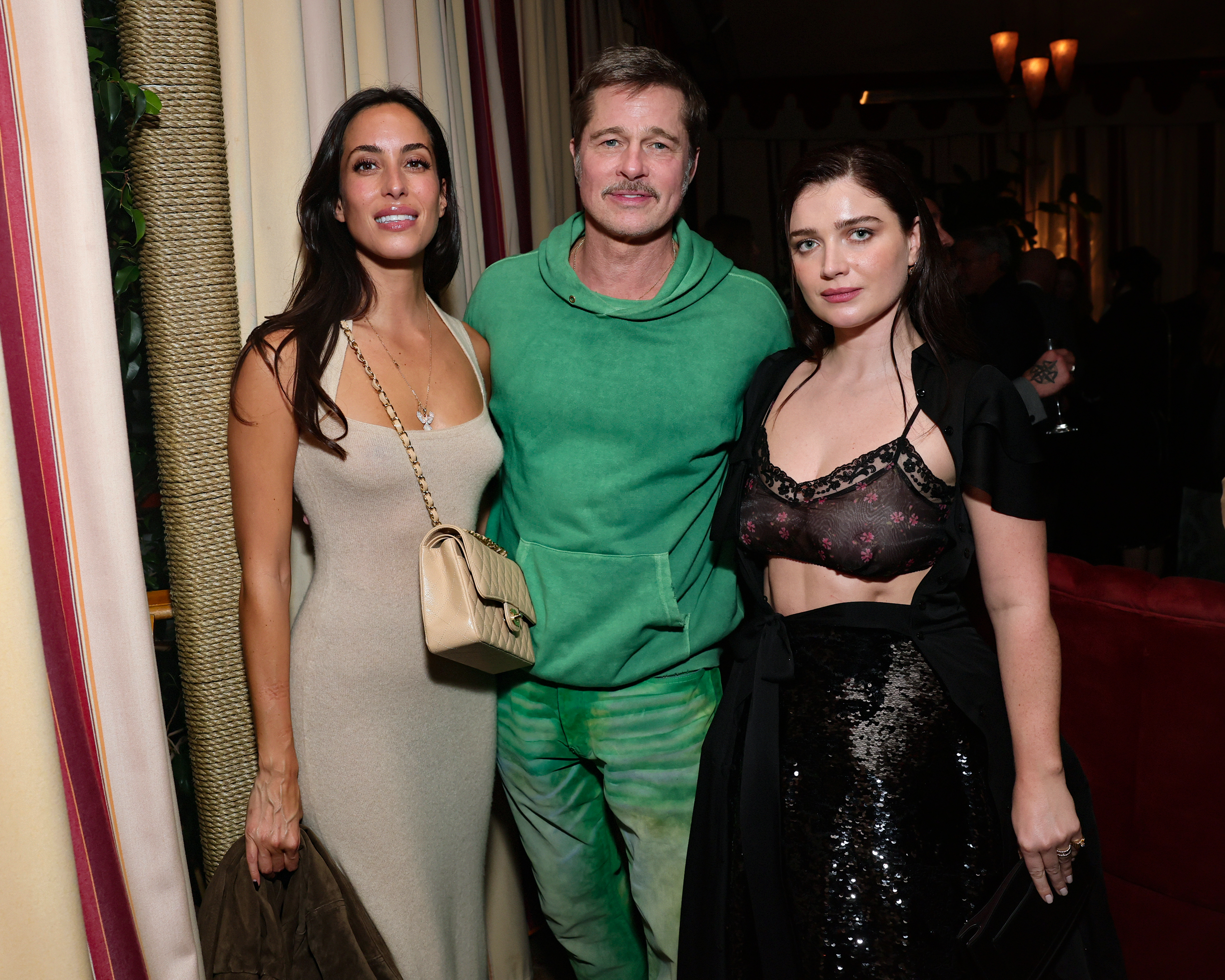 Ines de Ramon in a beige dress, Brad Pitt in a green outfit, and Eve Hewson in a black sequin skirt pose together at an indoor event.