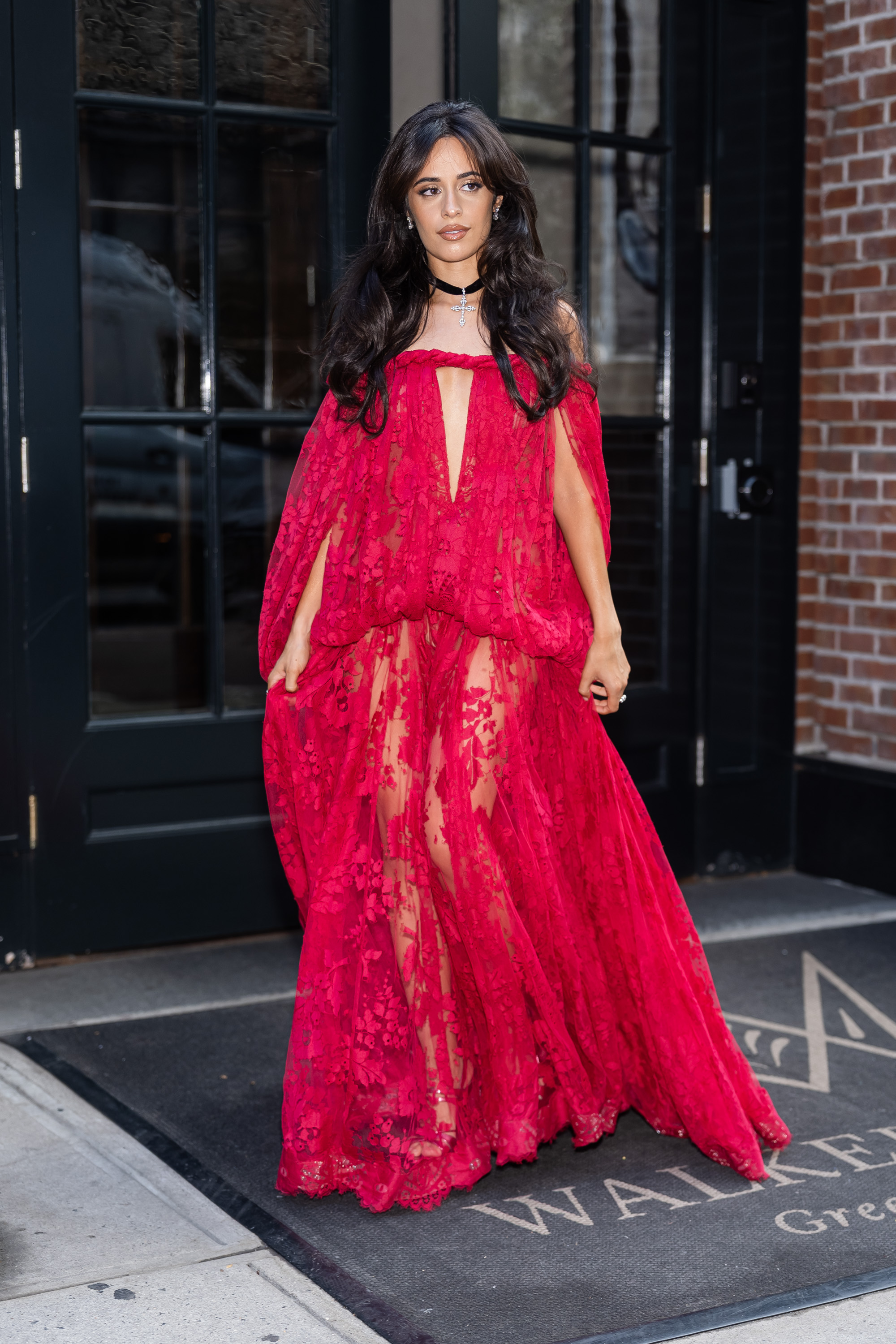 Camila Cabello wearing a floor-length red lace gown with a deep V-neckline and black choker, standing outside a building in New York City.
