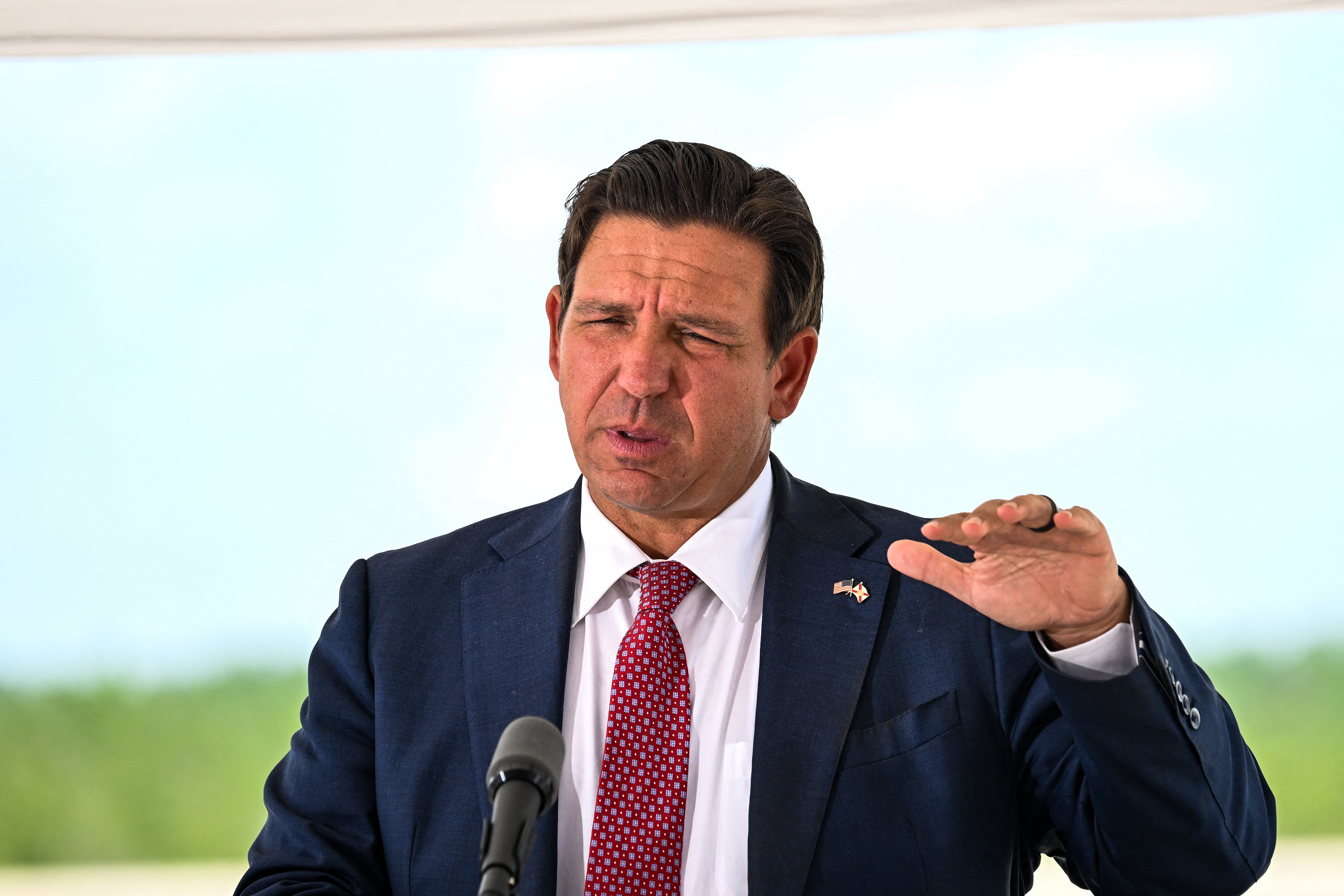 Ron DeSantis in a navy suit and red tie gesturing at a podium microphone during an outdoor press conference.