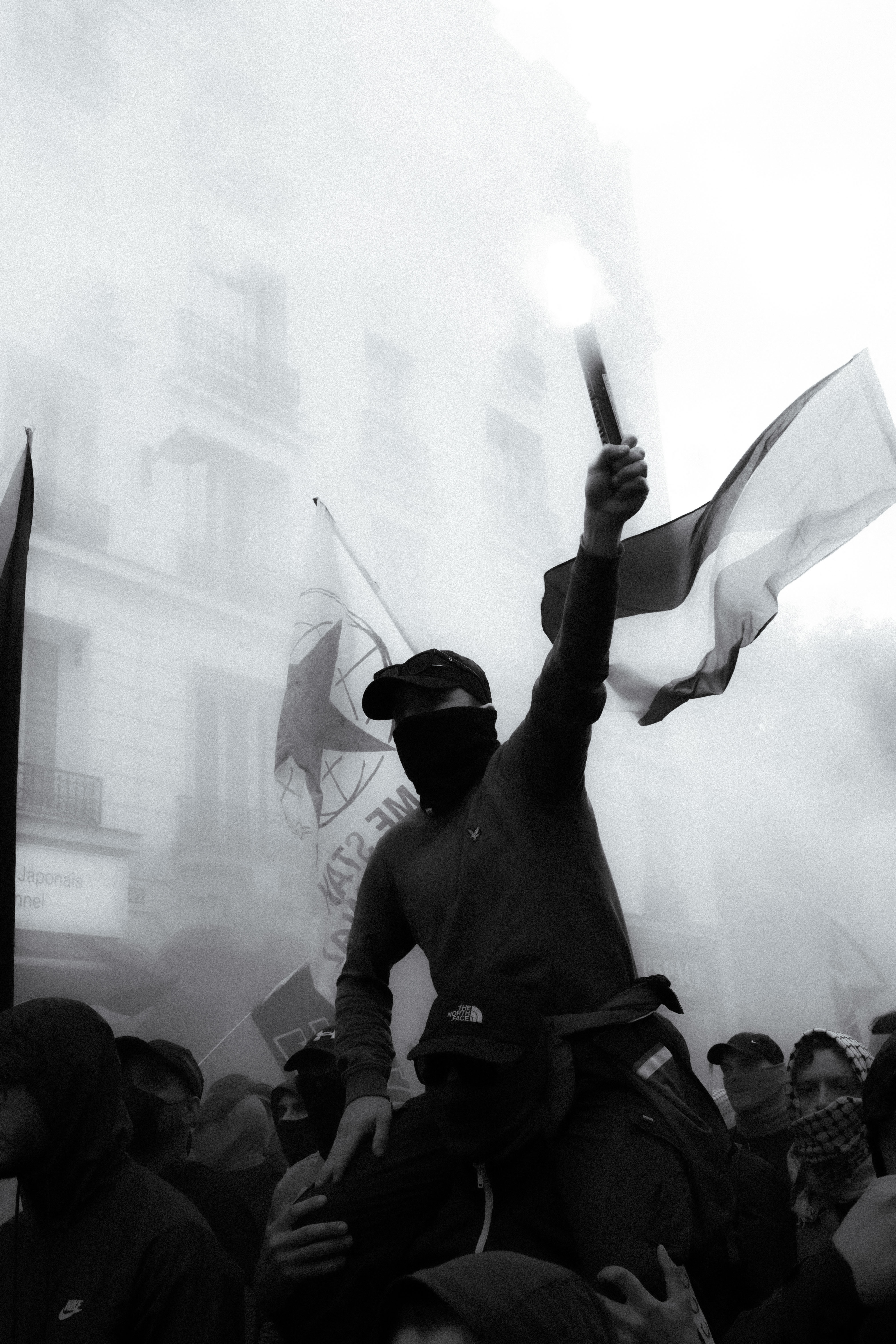 Masked protester holding a lit flare aloft amid smoke and flags at an Antifa demonstration in Paris.