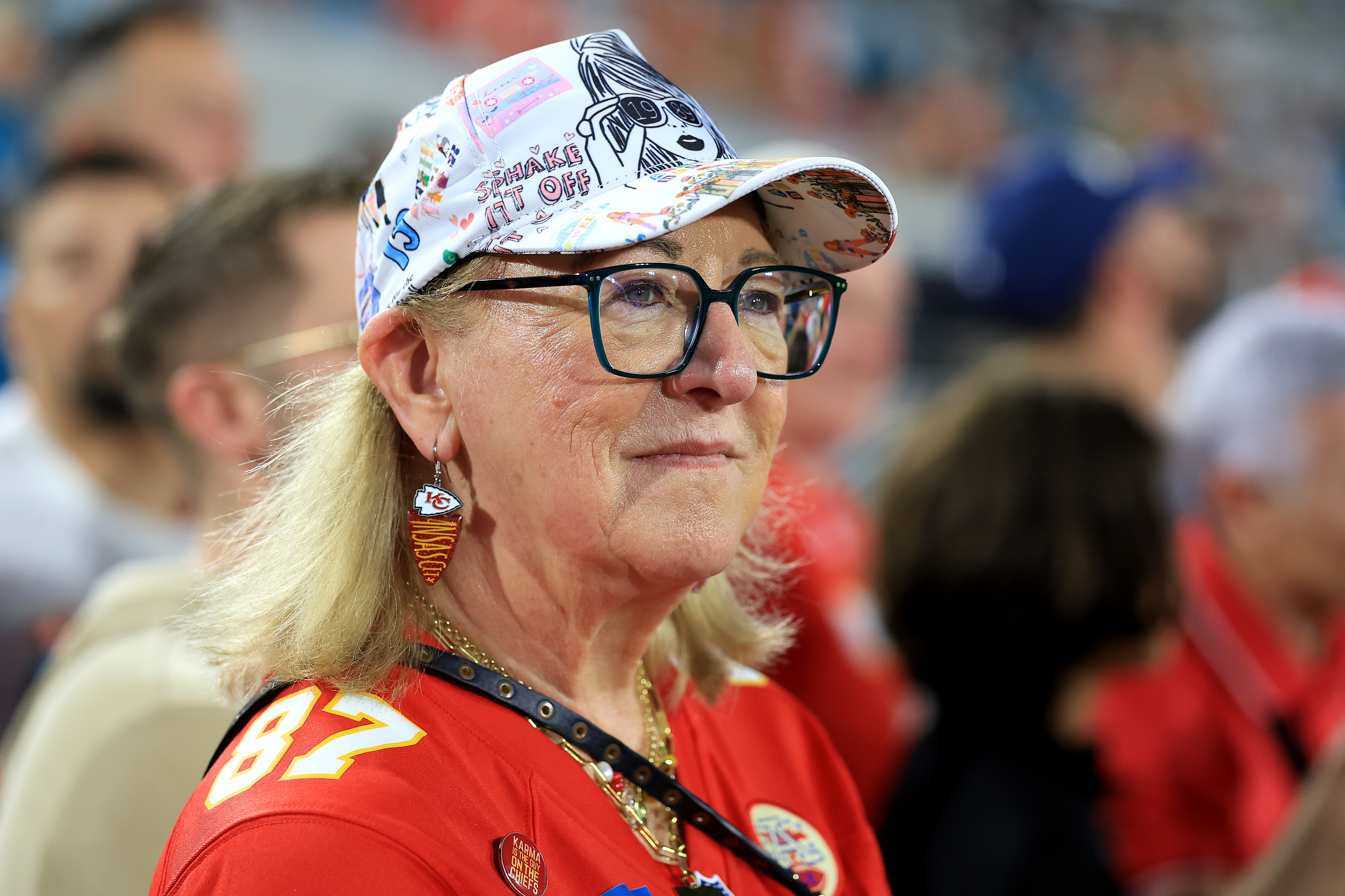 Donna Kelce in glasses, a Kansas City Chiefs No. 87 jersey, and a Taylor Swift-themed cap sitting in stadium stands.