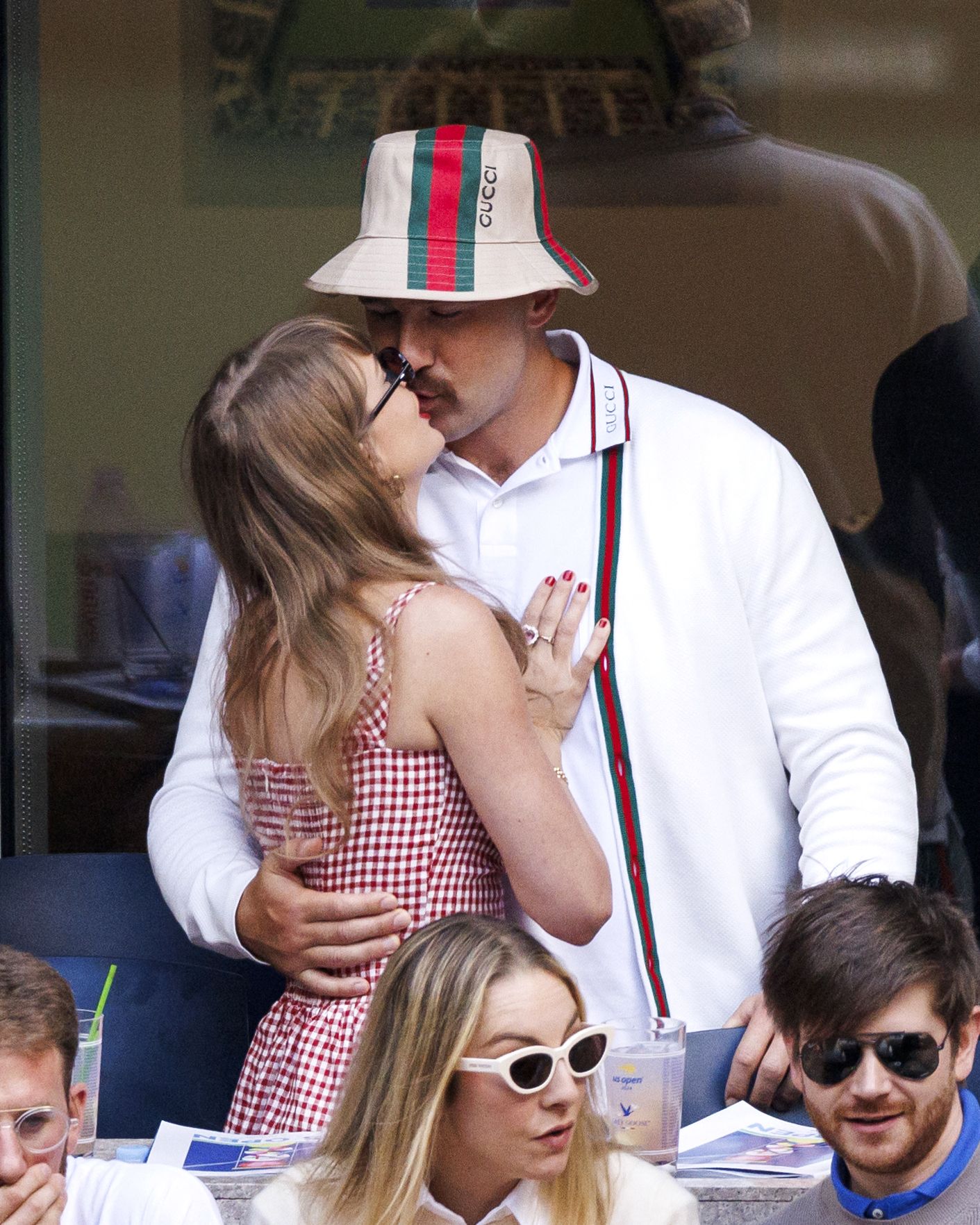 A couple kissing and embracing in the stands at the US Open, the man wearing a Gucci bucket hat and white polo, the woman in a red gingham dress.