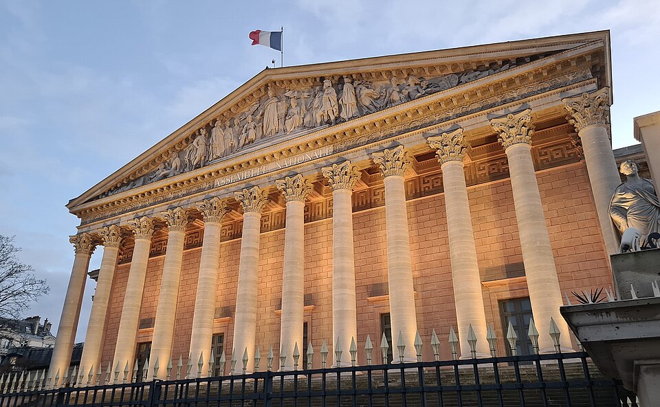 The French National Assembly building in Paris features classical architecture with prominent columns and intricate sculptures, illuminated against a twilight sky.