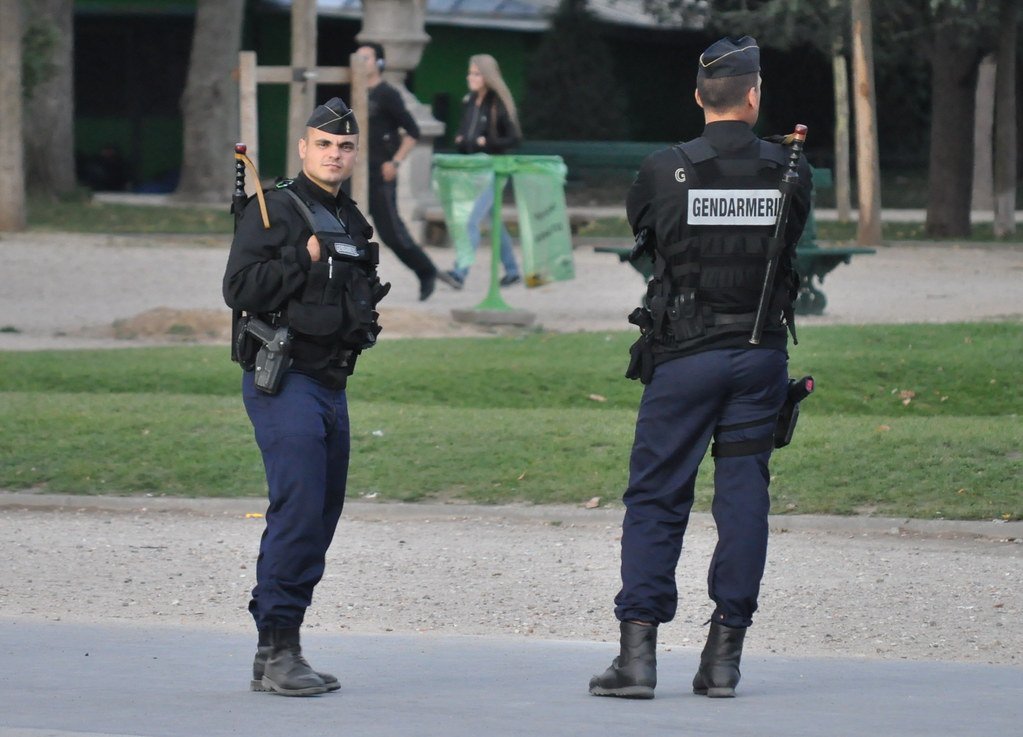 Two uniformed gendarmes stand in a park, one facing forward and the other turned away, showcasing their tactical gear and presence in a public space.