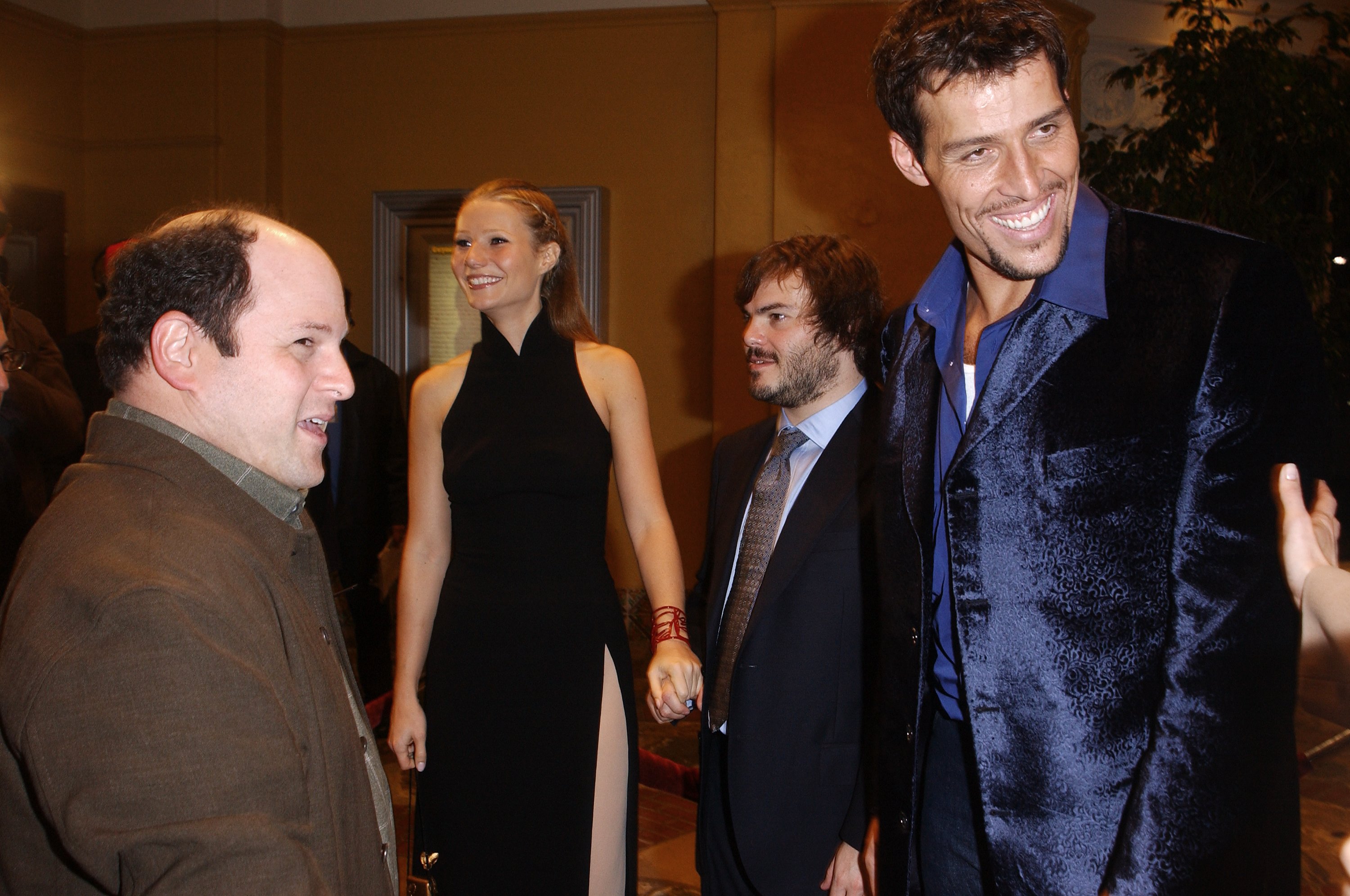 Four people — a bald man in brown, a tall blonde woman in black, a shorter bearded man in a suit, and a tall man in a blue satin jacket — chat at a film premiere.