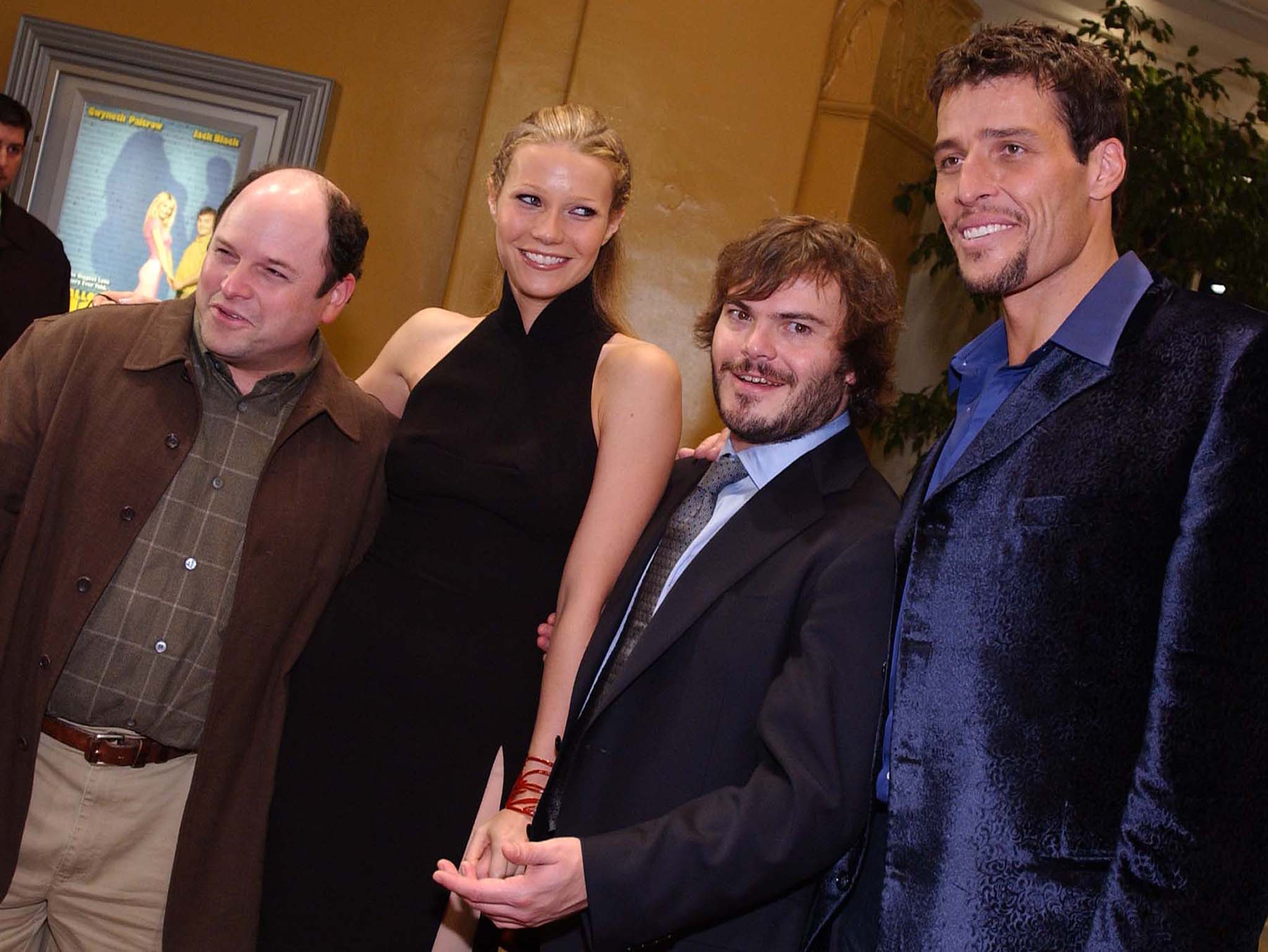 Four people posing at a movie premiere: a bald man in brown coat, a tall blonde woman in black dress, a shorter bearded man in a suit and tie, and a tall man in a blue blazer.