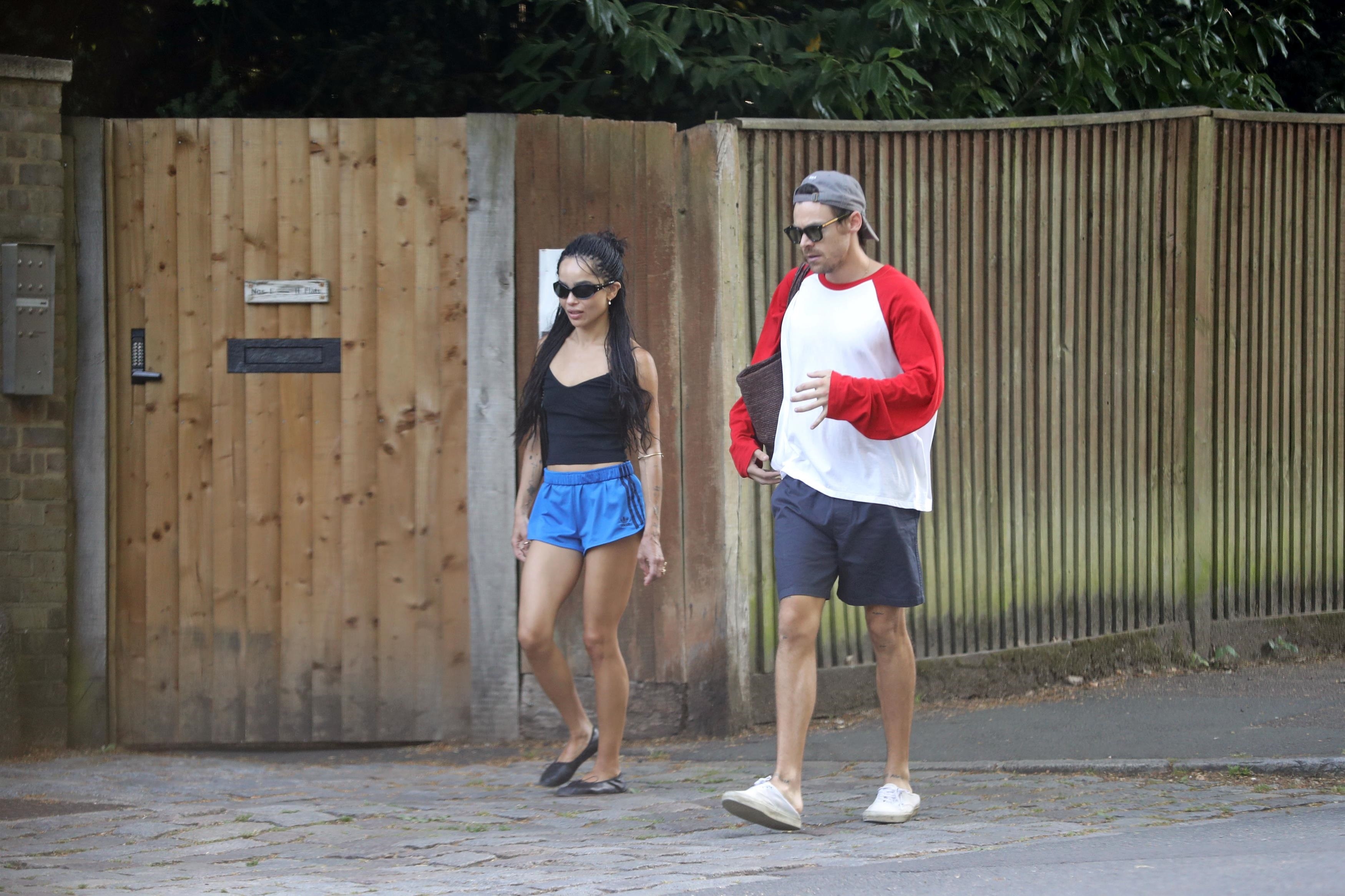 A woman in a black crop top and blue shorts walks alongside a man in a red and white baseball tee and navy shorts near a wooden fence.