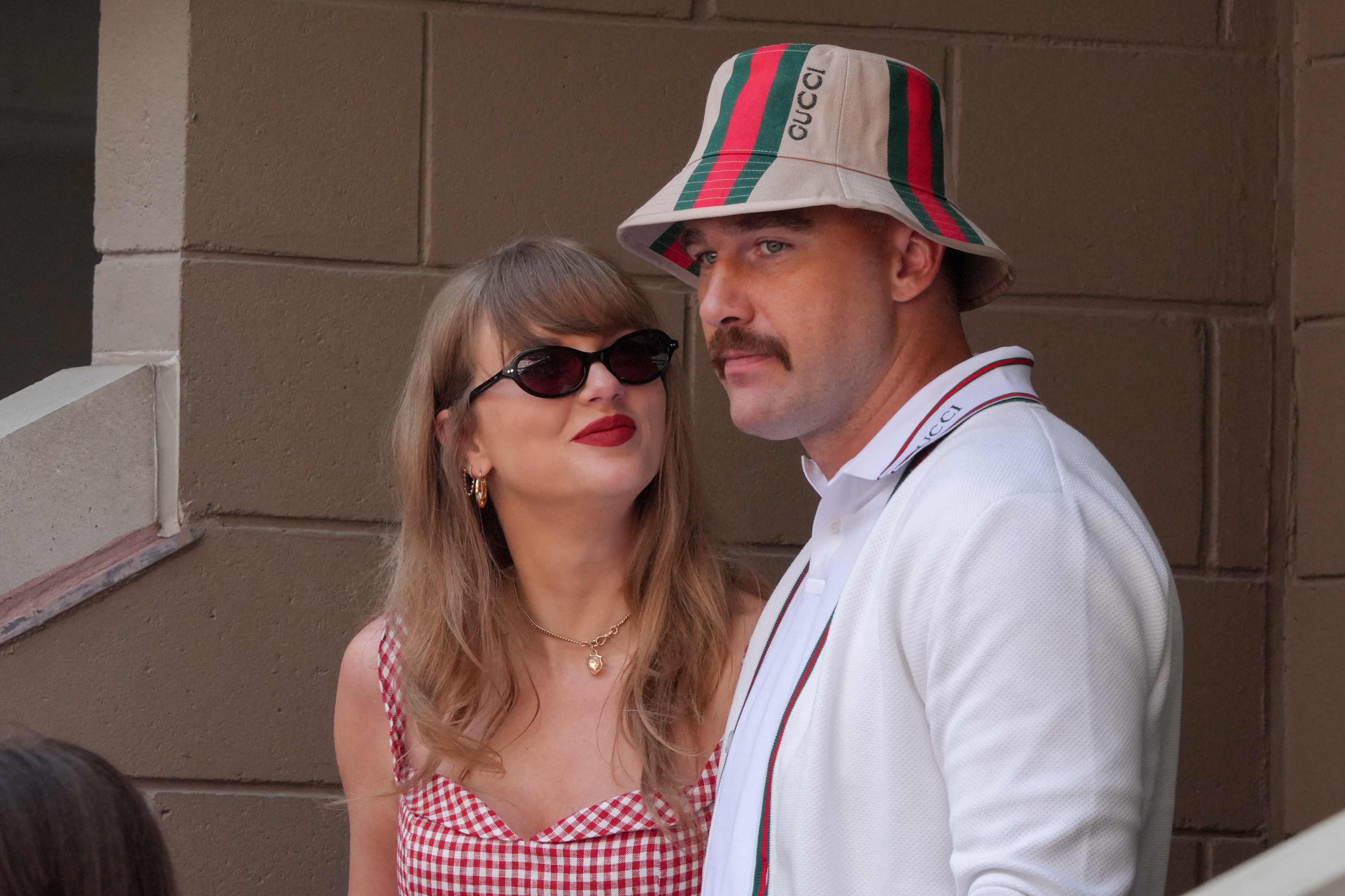 A woman in a red gingham dress with sunglasses and red lipstick stands beside a man wearing a Gucci bucket hat and white polo shirt against a brick wall backdrop.