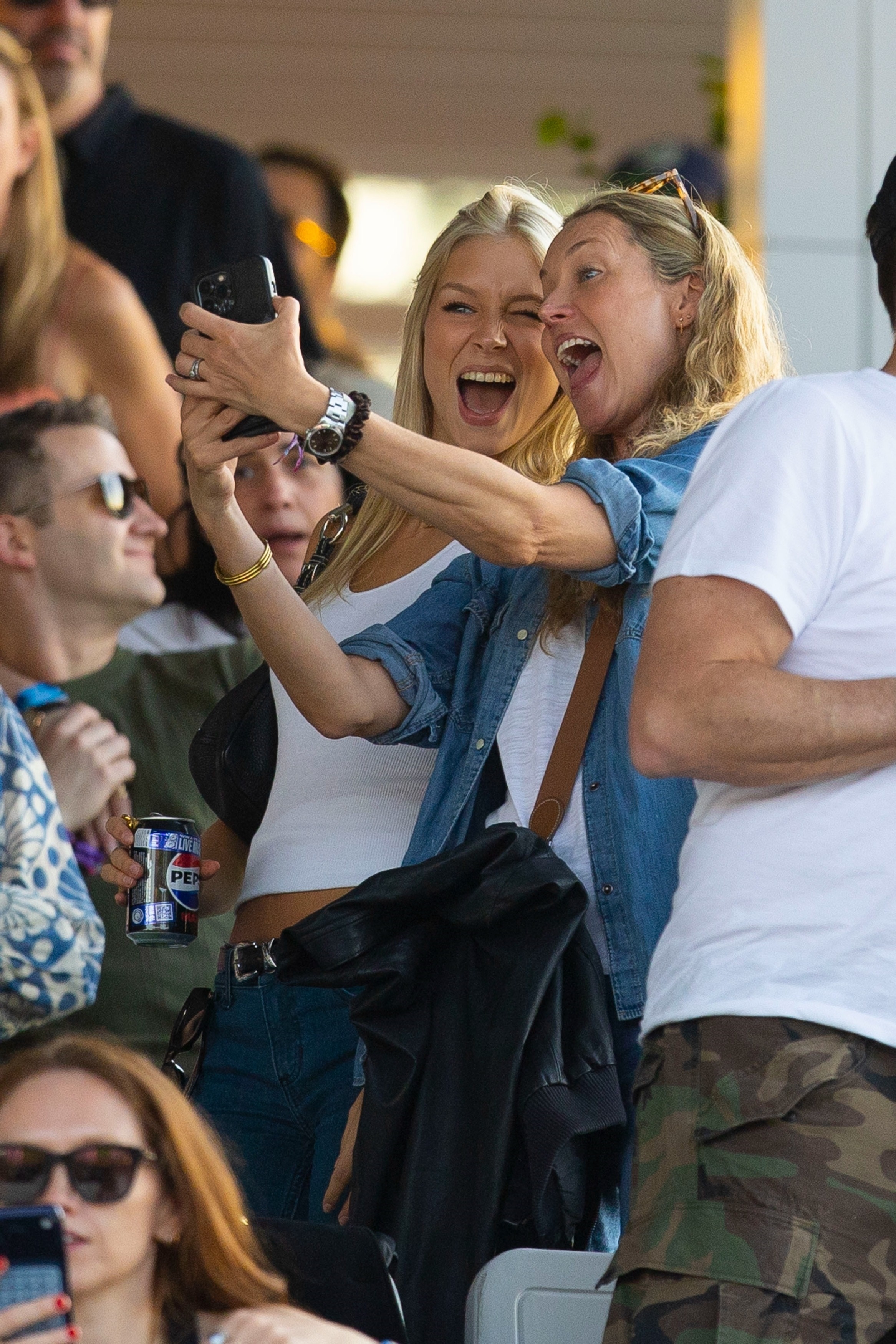 Two women laughing and taking a selfie together in a crowd at an outdoor concert, one in a white tank top and jeans.