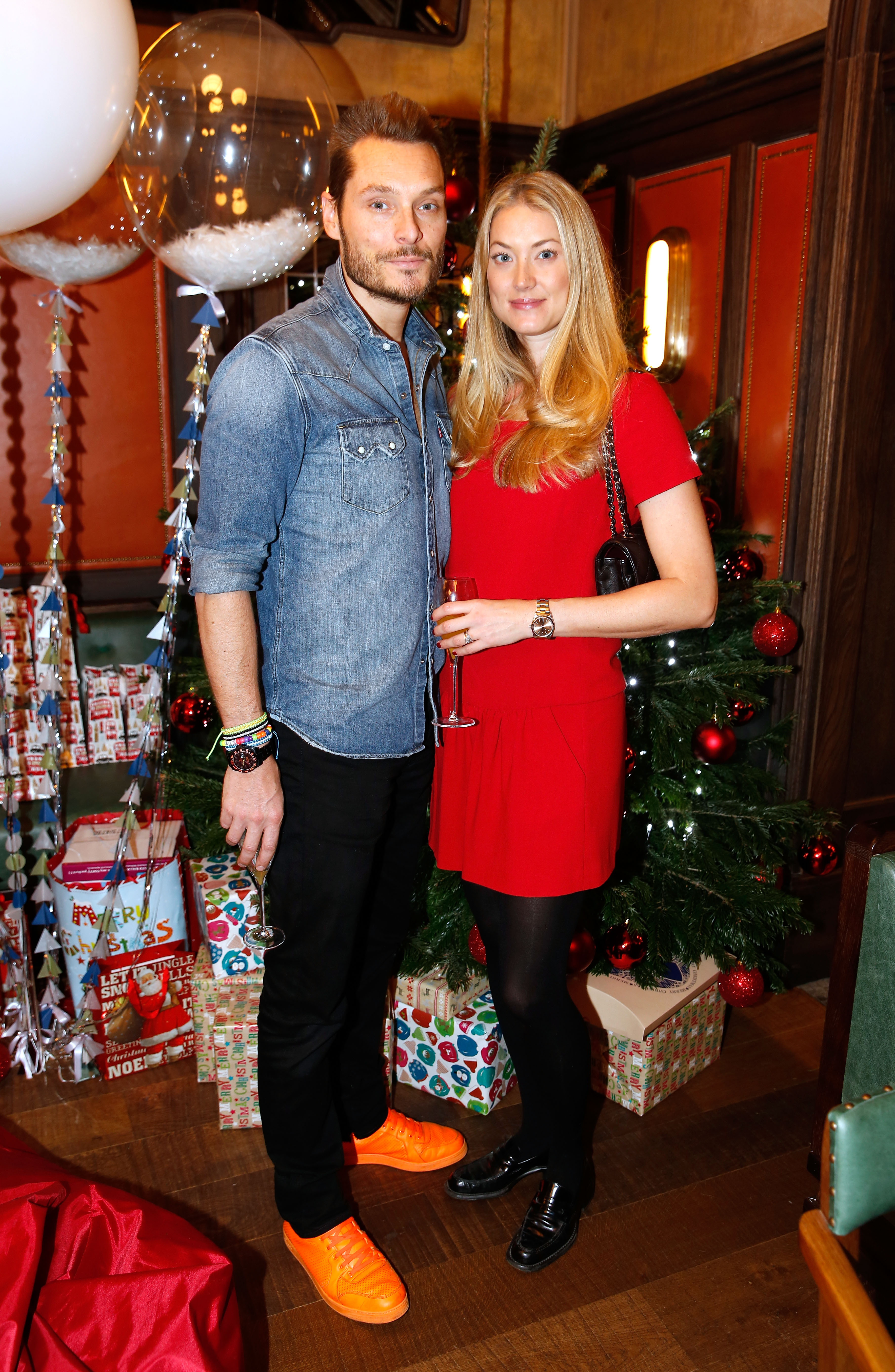 A man in a denim shirt and orange sneakers stands beside a blonde woman in a red dress holding a champagne flute, in front of a decorated Christmas tree.