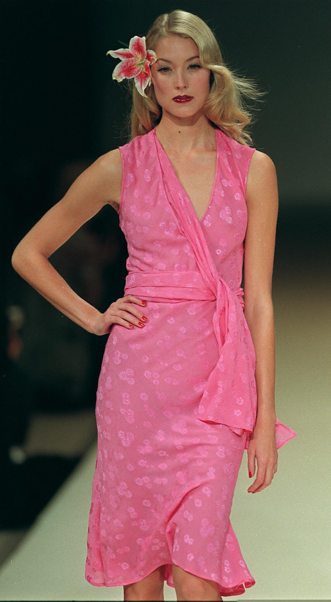 Blonde female model on a runway wearing a pink wrap dress with a floral lily hair accessory.