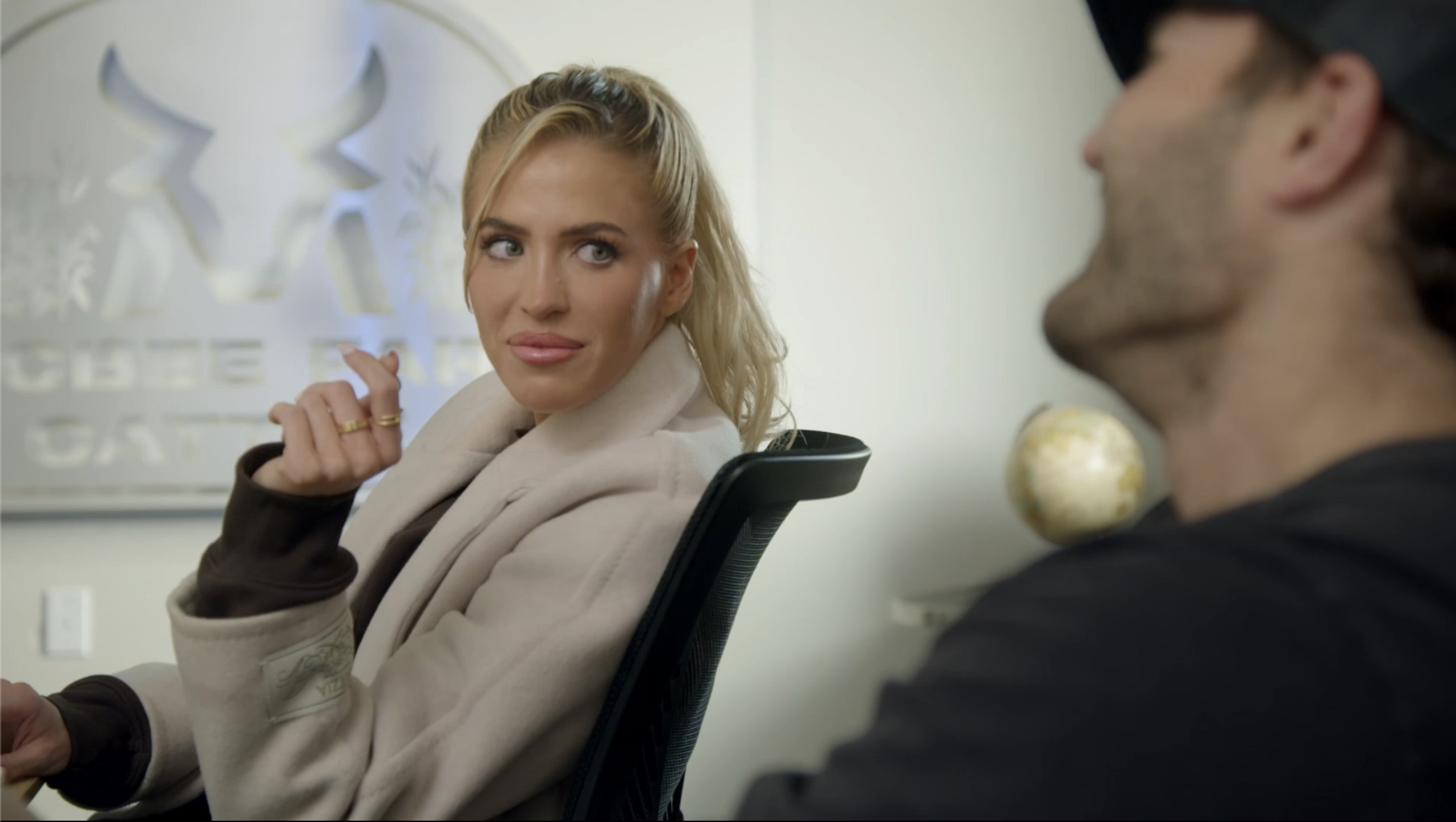 Blonde woman in a cream coat seated in an office chair, looking sideways at someone off-camera, with a logo on the wall behind her.