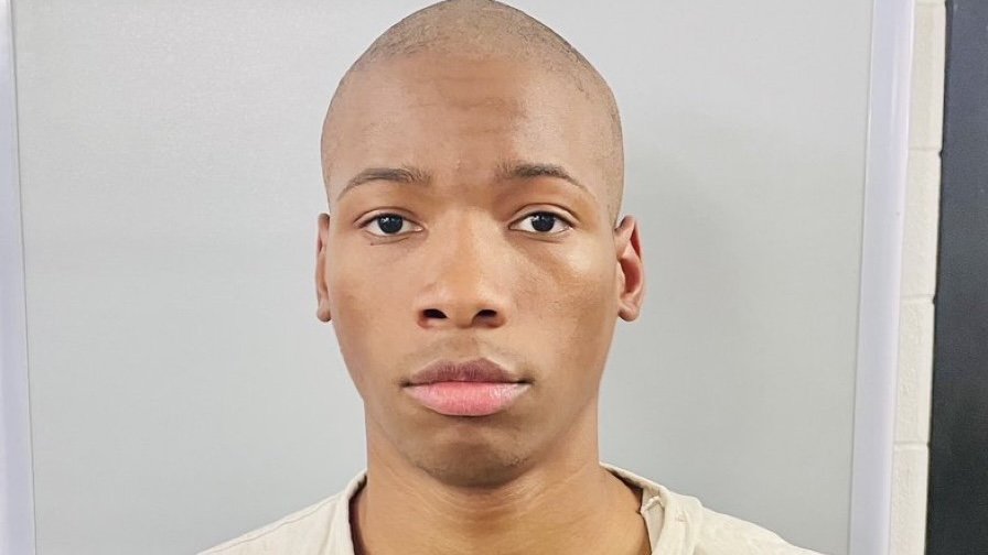 Young man with a shaved head and neutral expression, wearing a light-colored shirt, standing against a plain background.