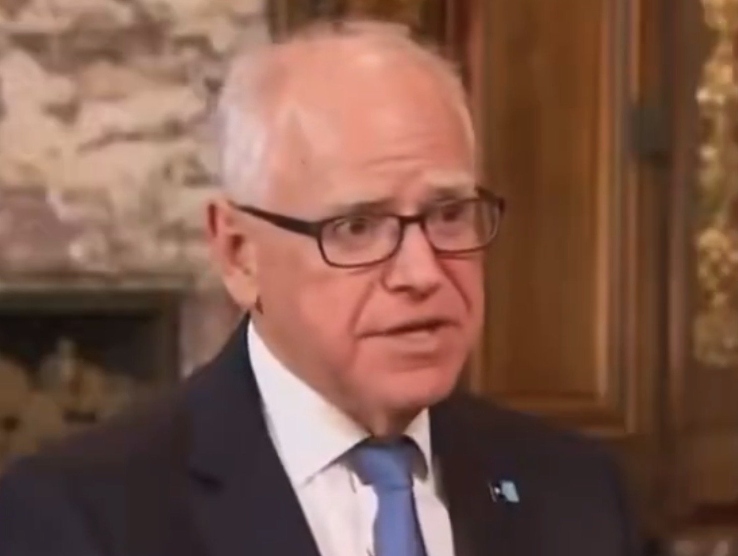 Business professional speaking in an elegant setting, wearing glasses and a suit with a blue tie, engaged in a serious conversation.